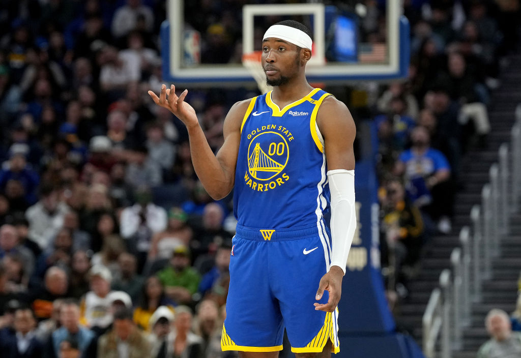 A basketball player in a Golden State Warriors uniform is on the court, gesturing with his hand. He's wearing a headband and arm sleeve, with a focused expression.