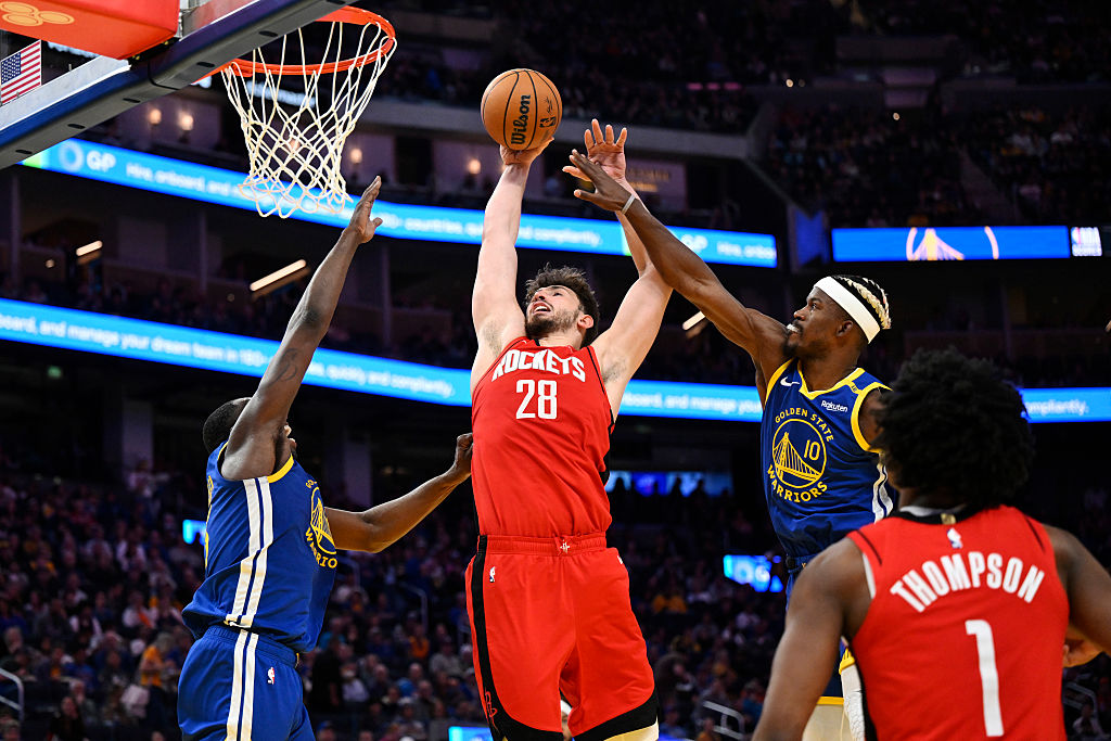 A basketball player in a red jersey leaps to shoot, surrounded by three opponents in blue jerseys. Two players attempt to block the shot near the hoop.