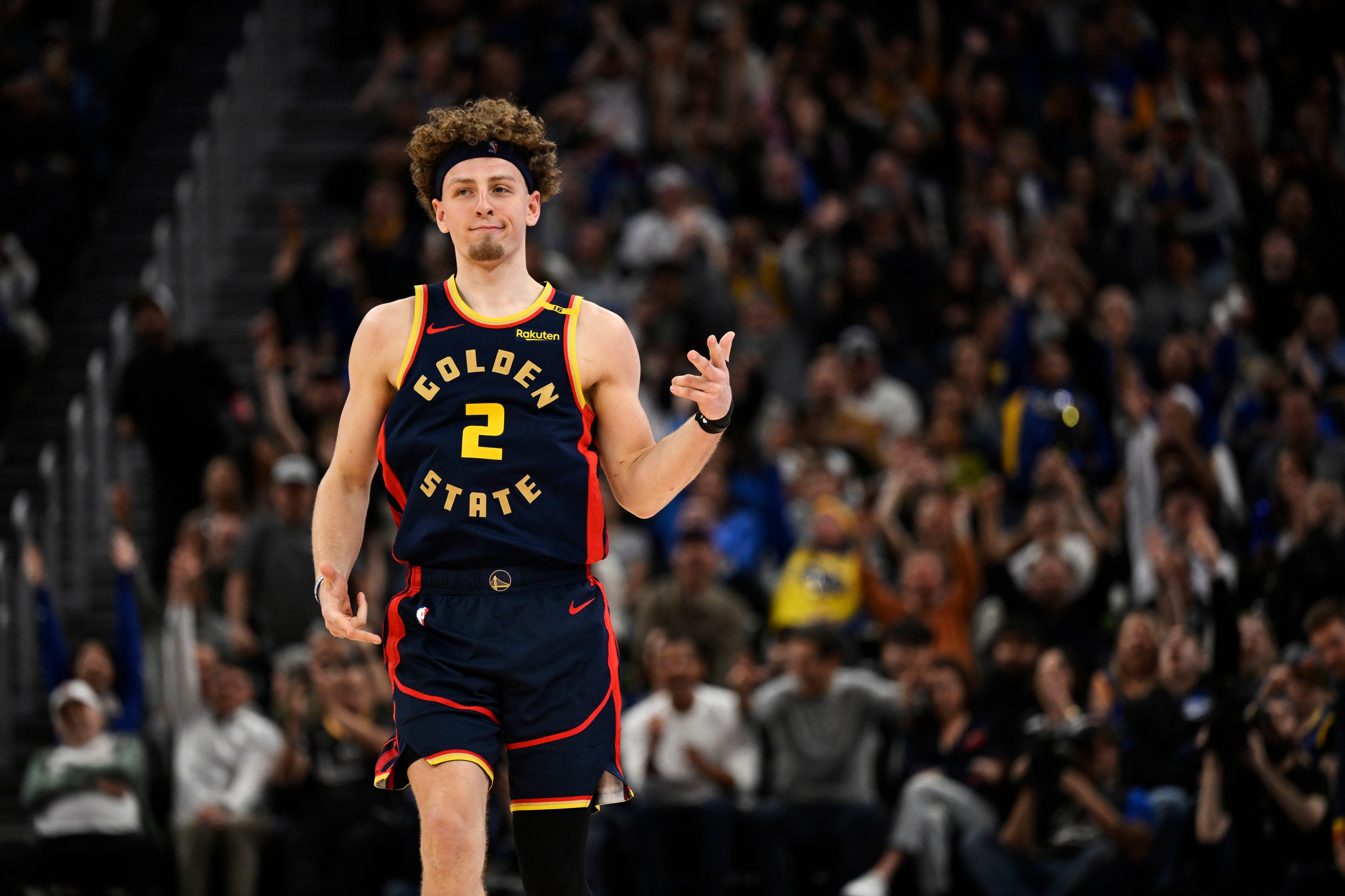 A basketball player wearing a Golden State jersey with the number 2 is making a hand gesture, possibly celebrating. A crowd in the background looks cheering.