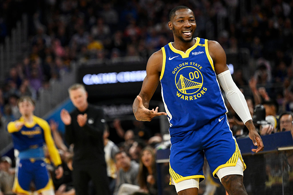 A basketball player in a Golden State Warriors uniform smiles on the court. The crowd is blurred in the background, and others on the sidelines cheer him on.