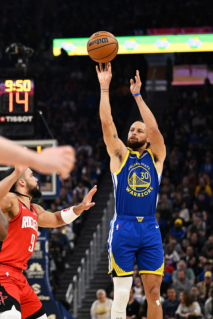 A basketball player in a blue Golden State Warriors jersey is shooting the ball mid-air. Another player in a red Rockets jersey is nearby. The scoreboard shows 9:50.