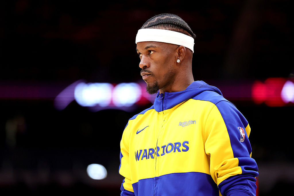 A basketball player in a yellow and blue Warriors jacket and a white headband stands focused, with braided hair. The background is blurred and dark.
