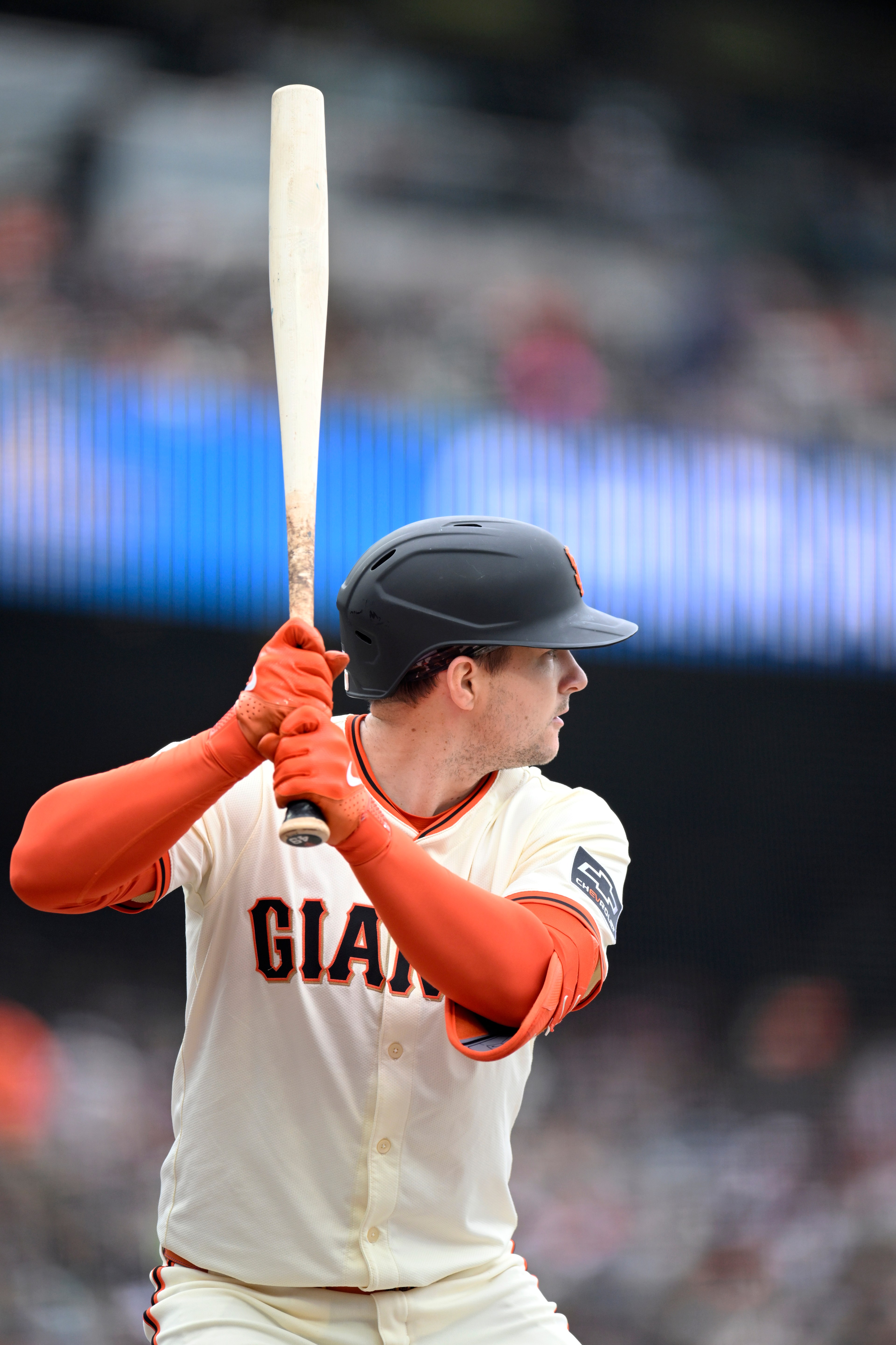 A baseball player in a white &quot;Giants&quot; jersey and black helmet is poised to bat, holding a light-colored bat, with a blurred stadium background.