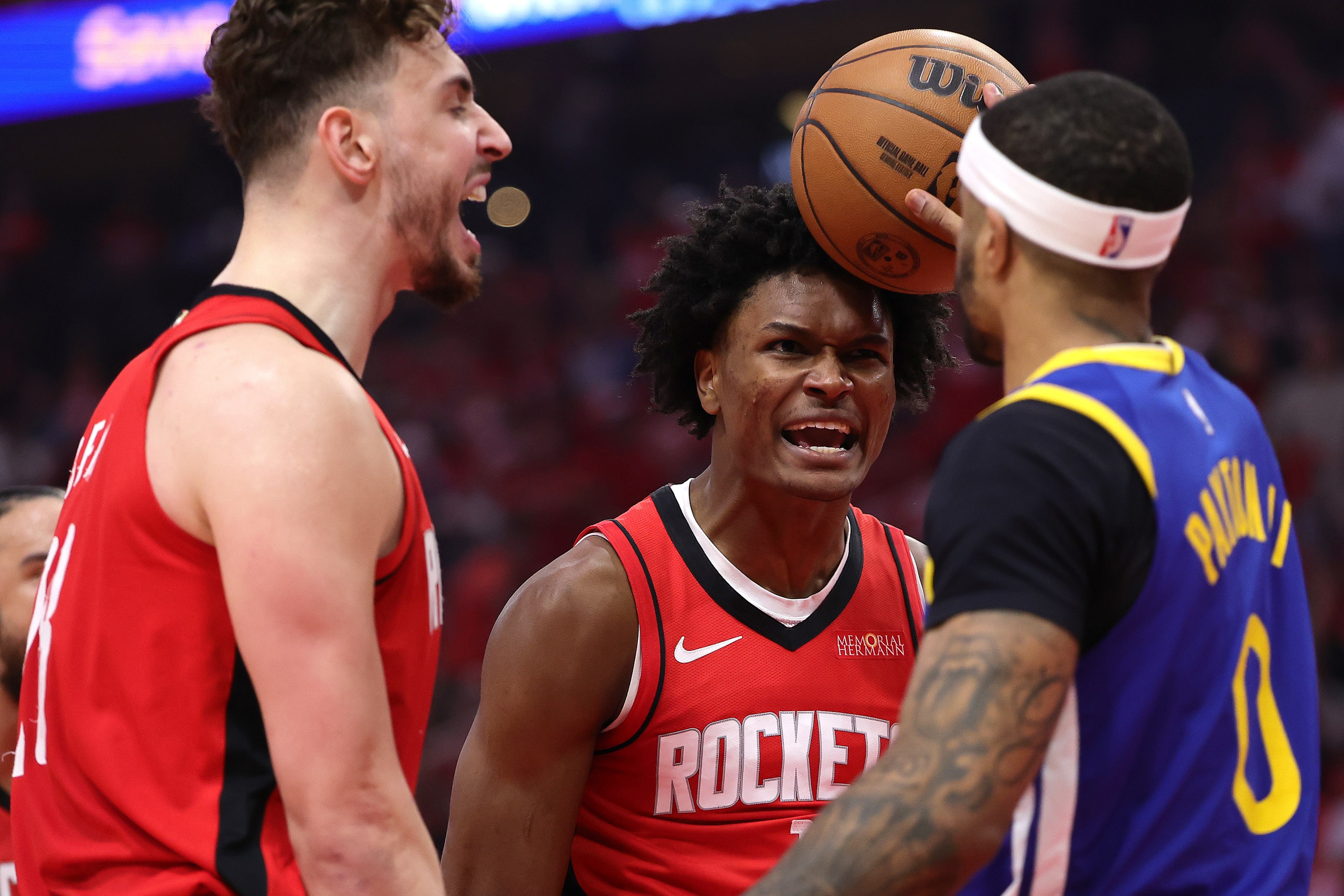 Three basketball players are on the court, two in red jerseys and one in blue, appearing tense. One player in red is yelling, the other holds a basketball.