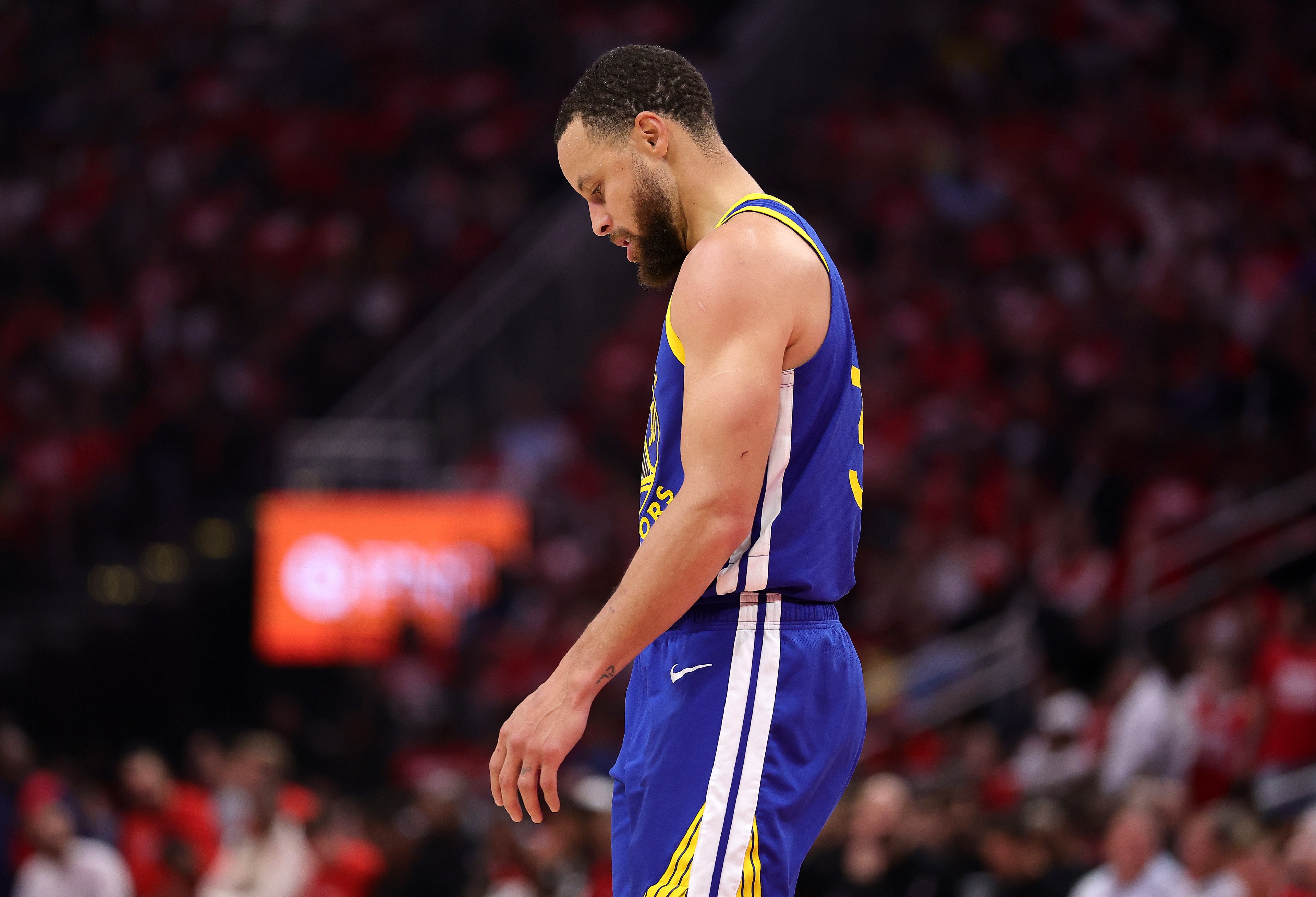 A basketball player in a blue and yellow jersey looks down, appearing contemplative, standing on the court with a blurred audience in the background.
