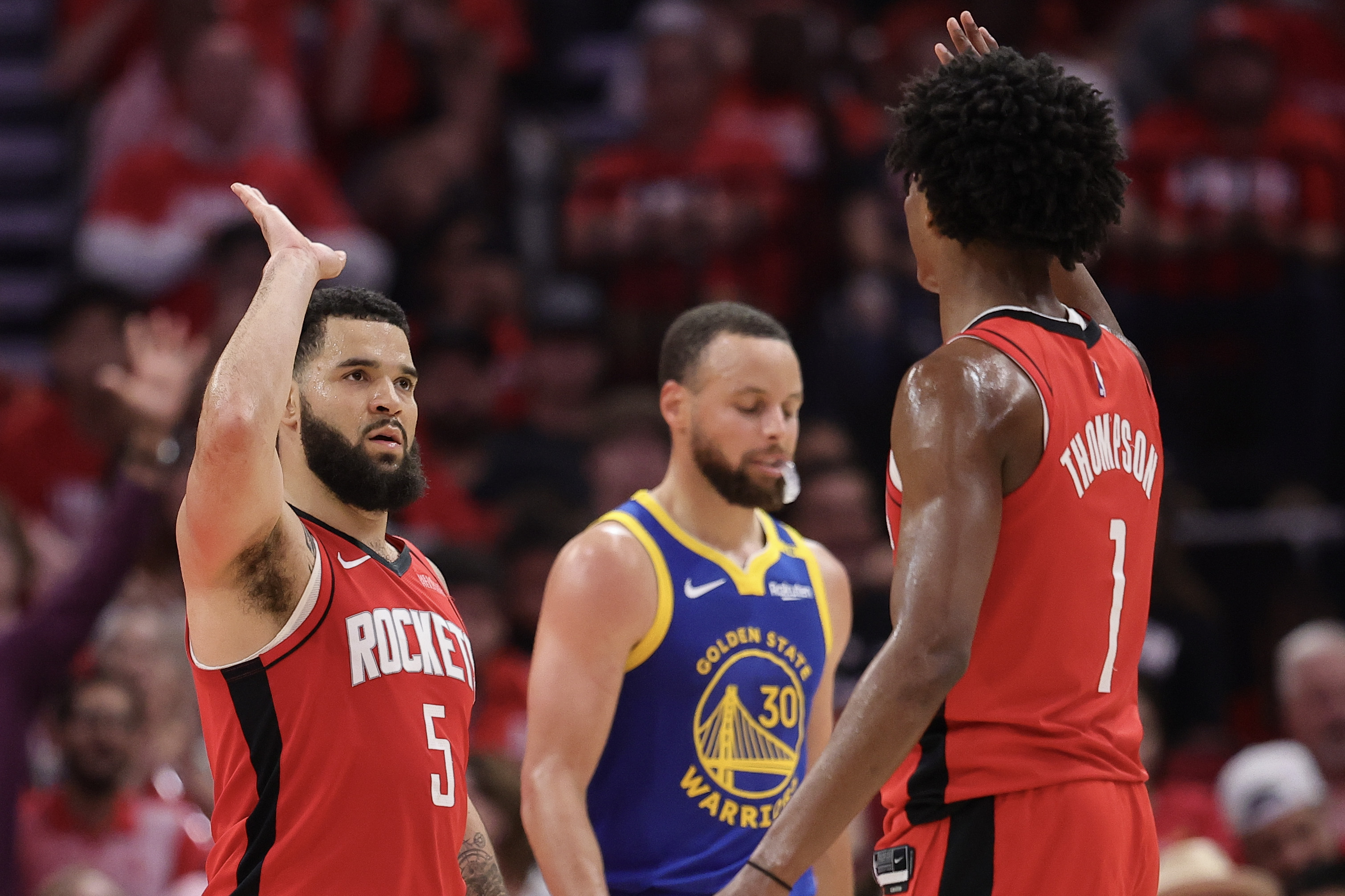 Two basketball players in red jerseys high-five, while a player in a blue jersey walks past them. The background shows a cheering crowd.
