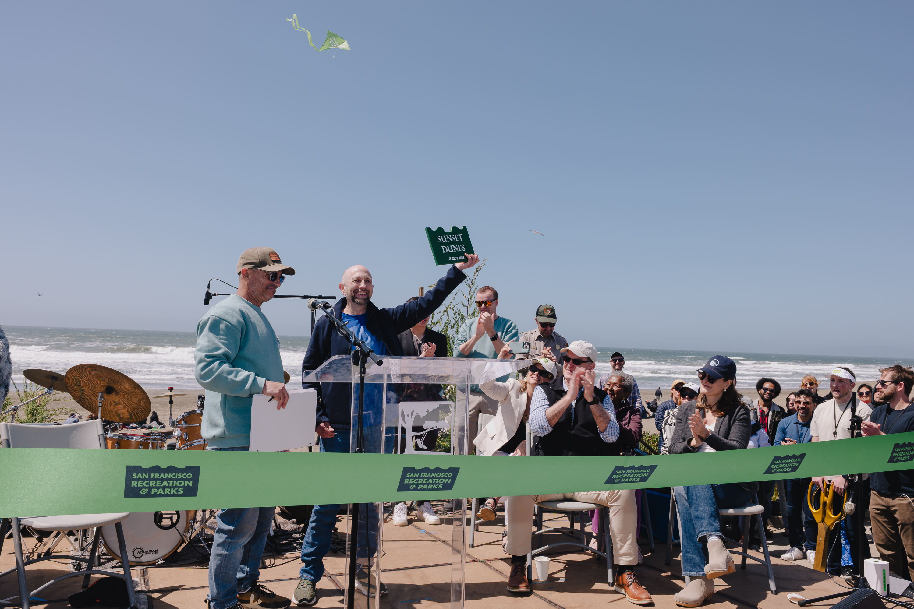People gather by the ocean, smiling and clapping, as a man holds up a “Sunset Dunes” sign. A band setup is nearby under a clear sky.