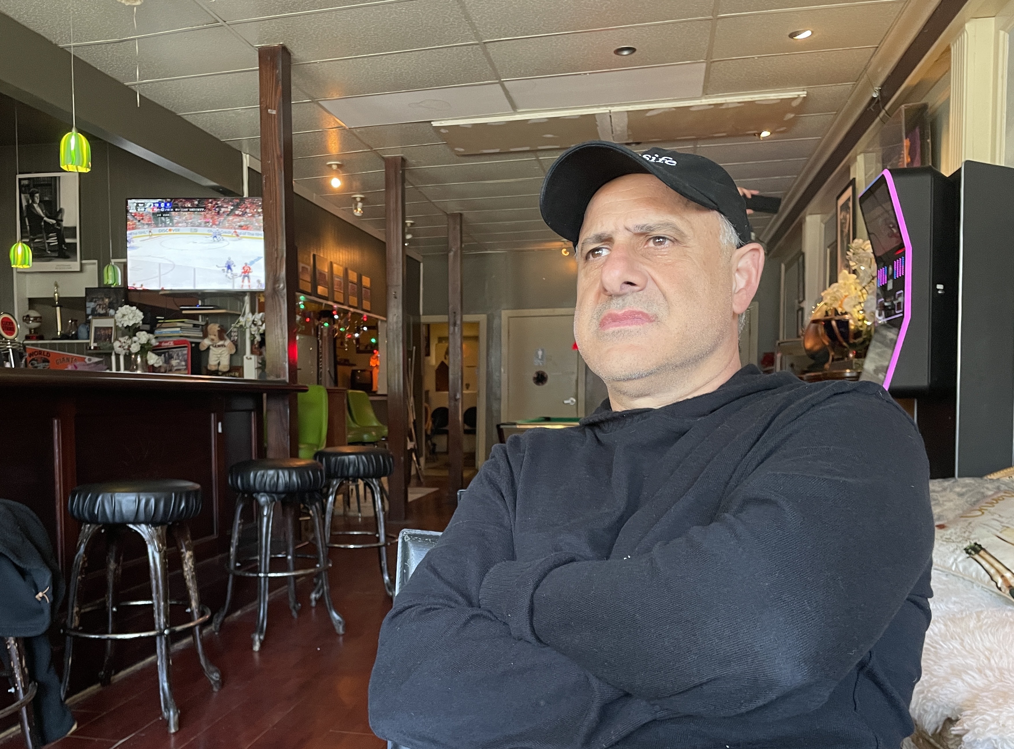 A man in a black cap and hoodie sits with arms crossed in a dimly lit café. Behind him, barstools and a TV displaying a hockey game are visible.
