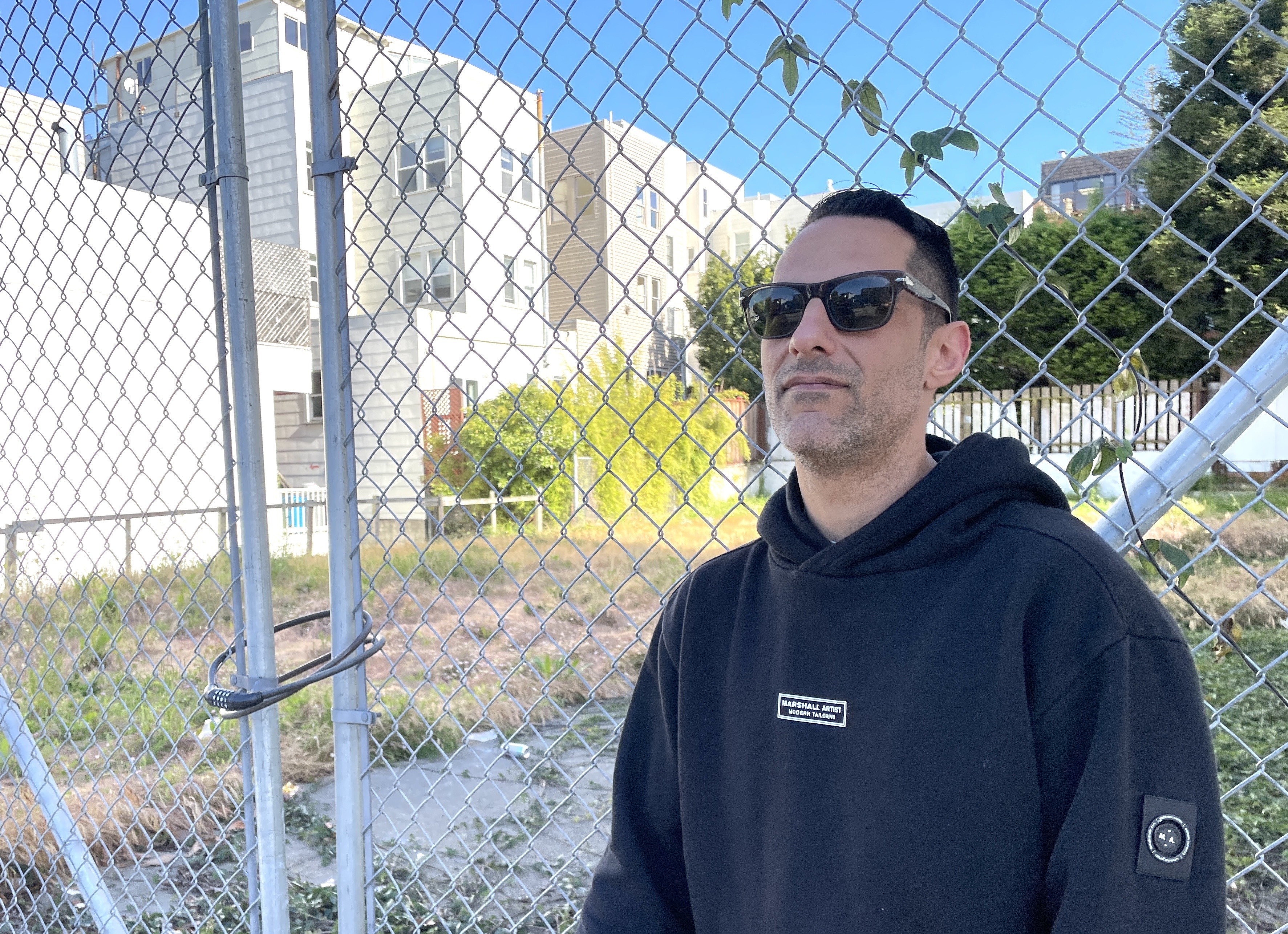 A person wearing sunglasses and a black hoodie stands in front of a chain-link fence. Behind them, there are residential buildings and greenery.