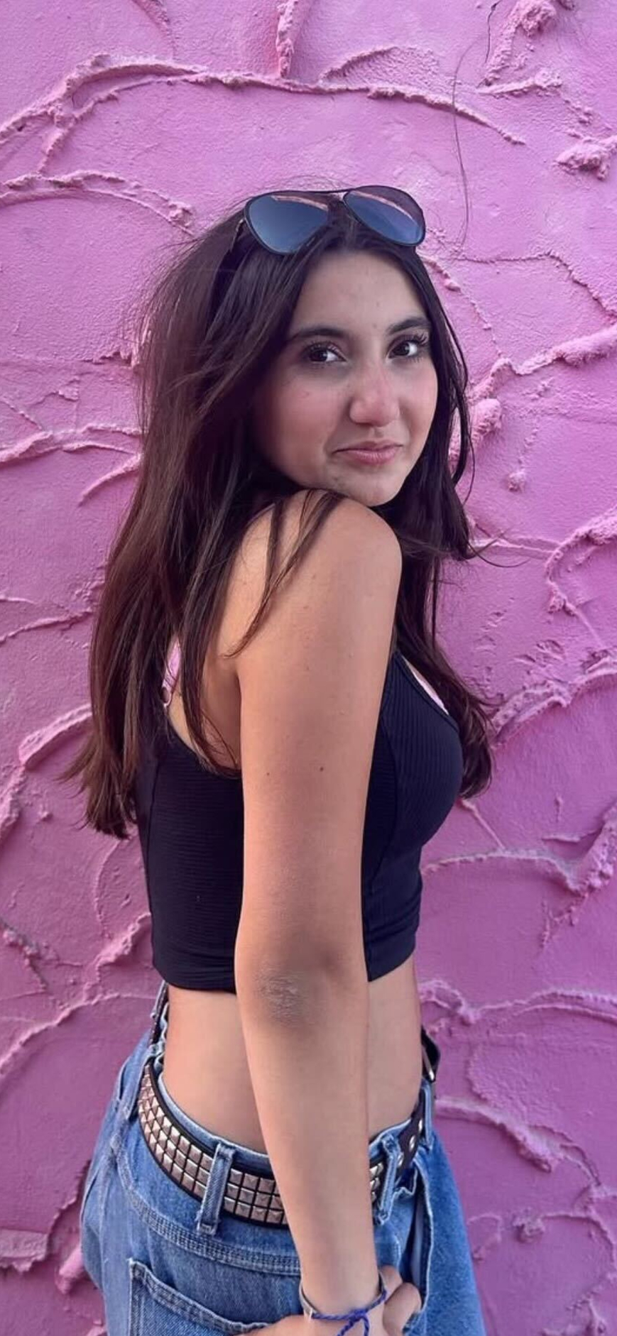 A girl with long hair and sunglasses on their head stands smiling in front of a textured pink wall. They wear a black top, jeans, and a studded belt.