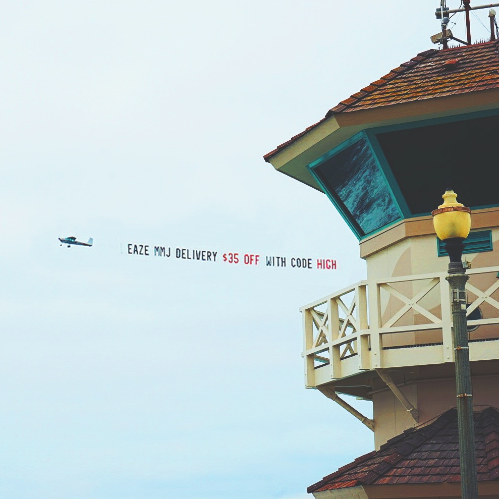 A small plane tows a banner reading, &quot;Eaze MMJ Delivery $35 off with code HIGH,&quot; near a tower with a wooden roof and a streetlight.