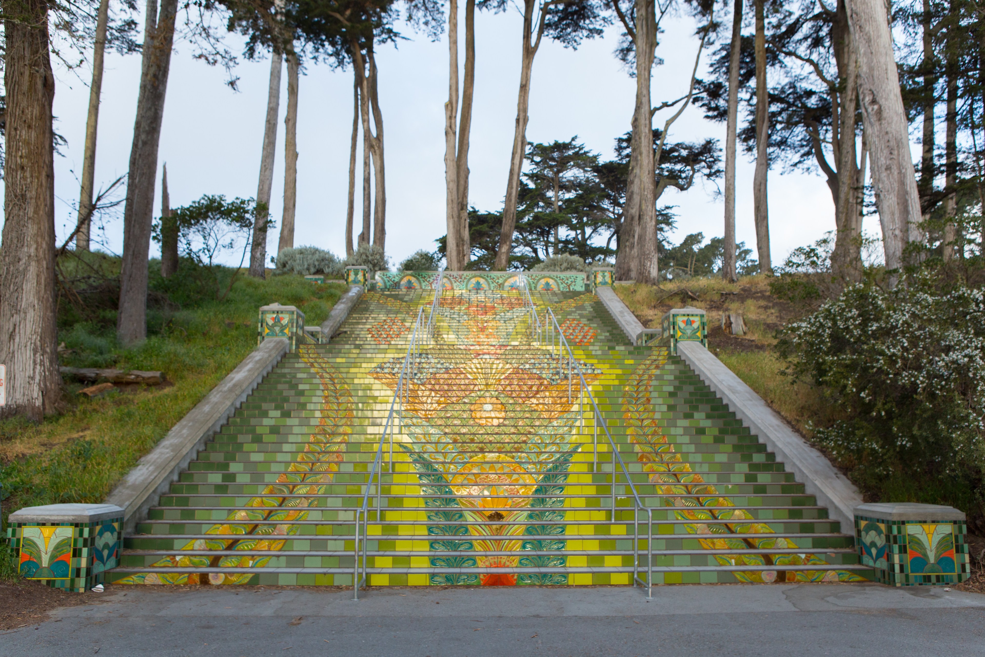 A colorful mosaic staircase with intricate patterns leads upward, flanked by greenery and tall trees, creating a vibrant, artistic pathway.