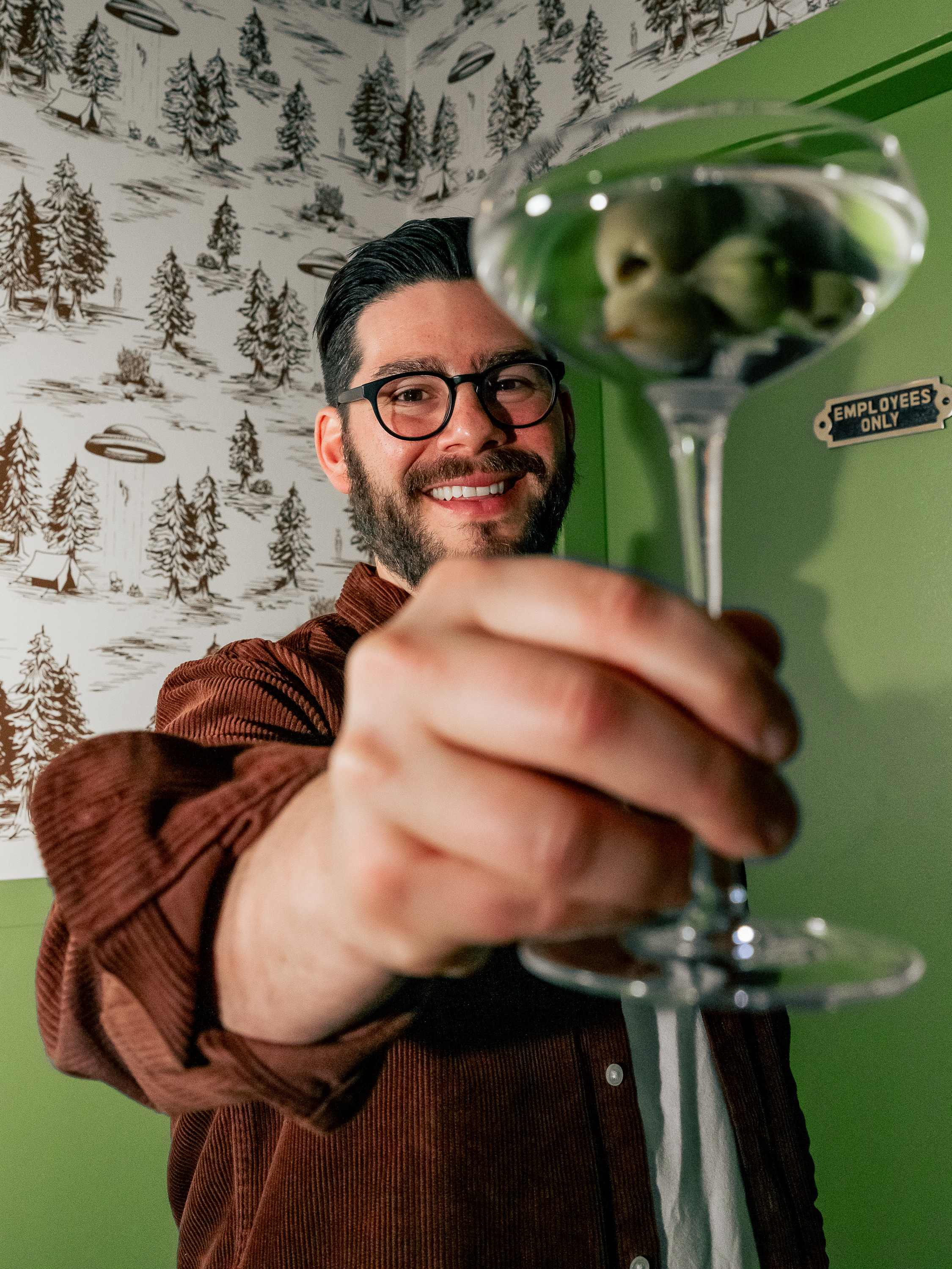 A person wearing glasses and a brown shirt smiles at the camera, holding up a martini glass with olives. The background features a forest-themed wallpaper.