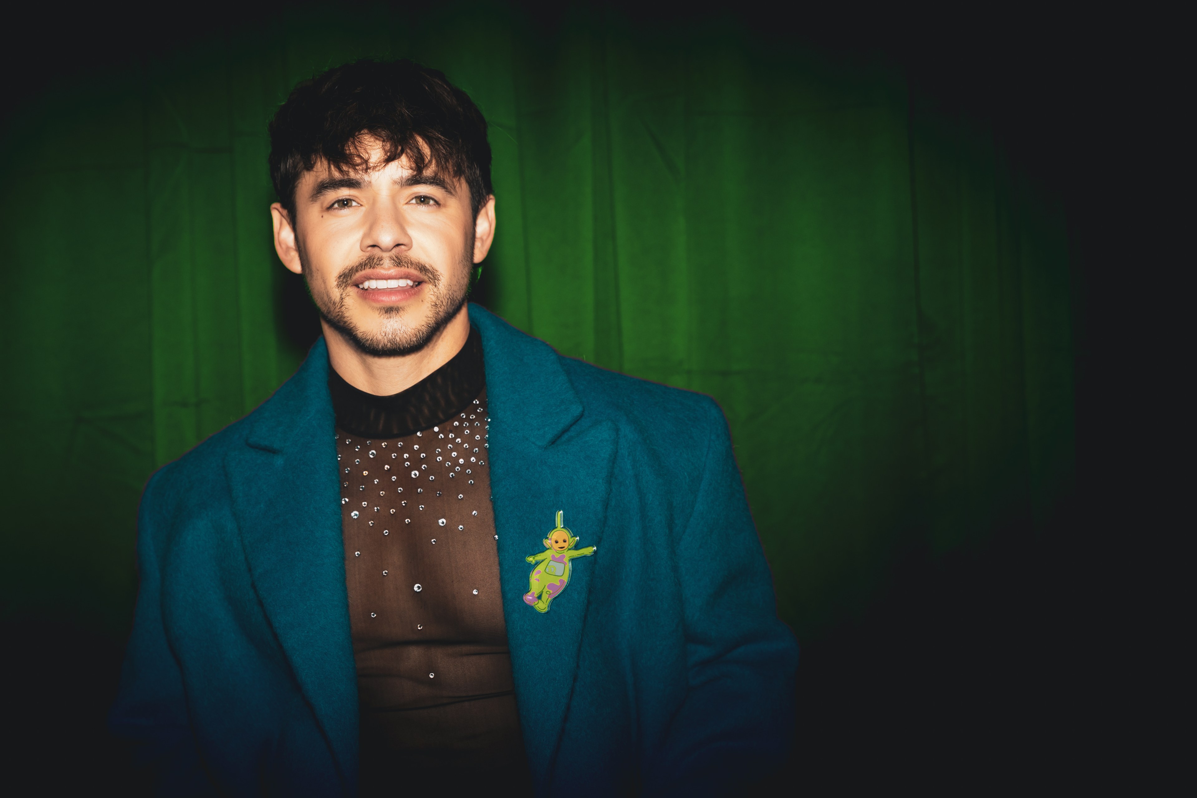 Singer David Archuleta stands against a dark green background, wearing a blue coat over a black, pearl-studded shirt. A colorful pin in the shape of an alien is on the coat.