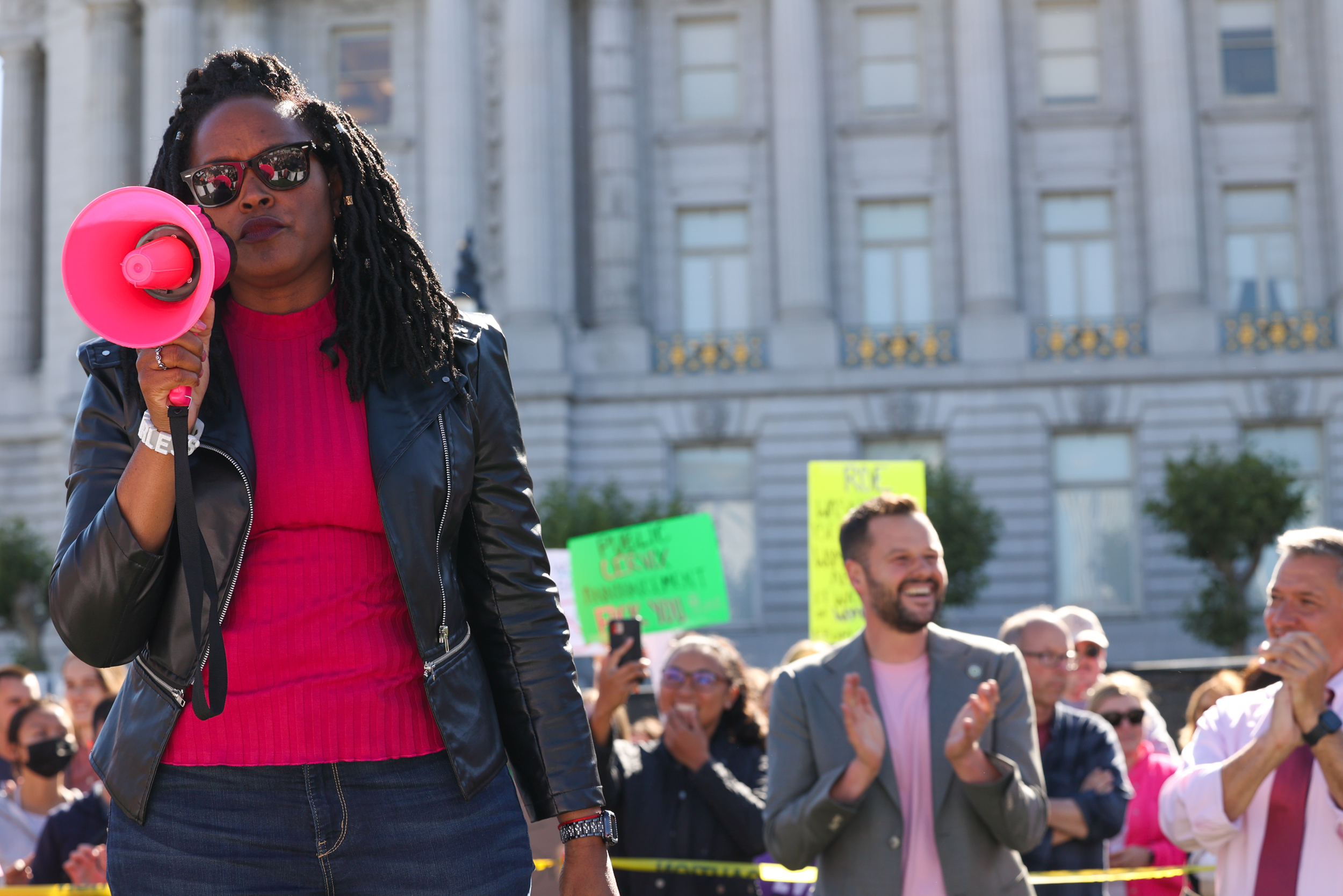 A woman with a pink bullhorn stands in front of a crowd. She wears sunglasses, a leather jacket, and a pink shirt. People in the background are clapping.