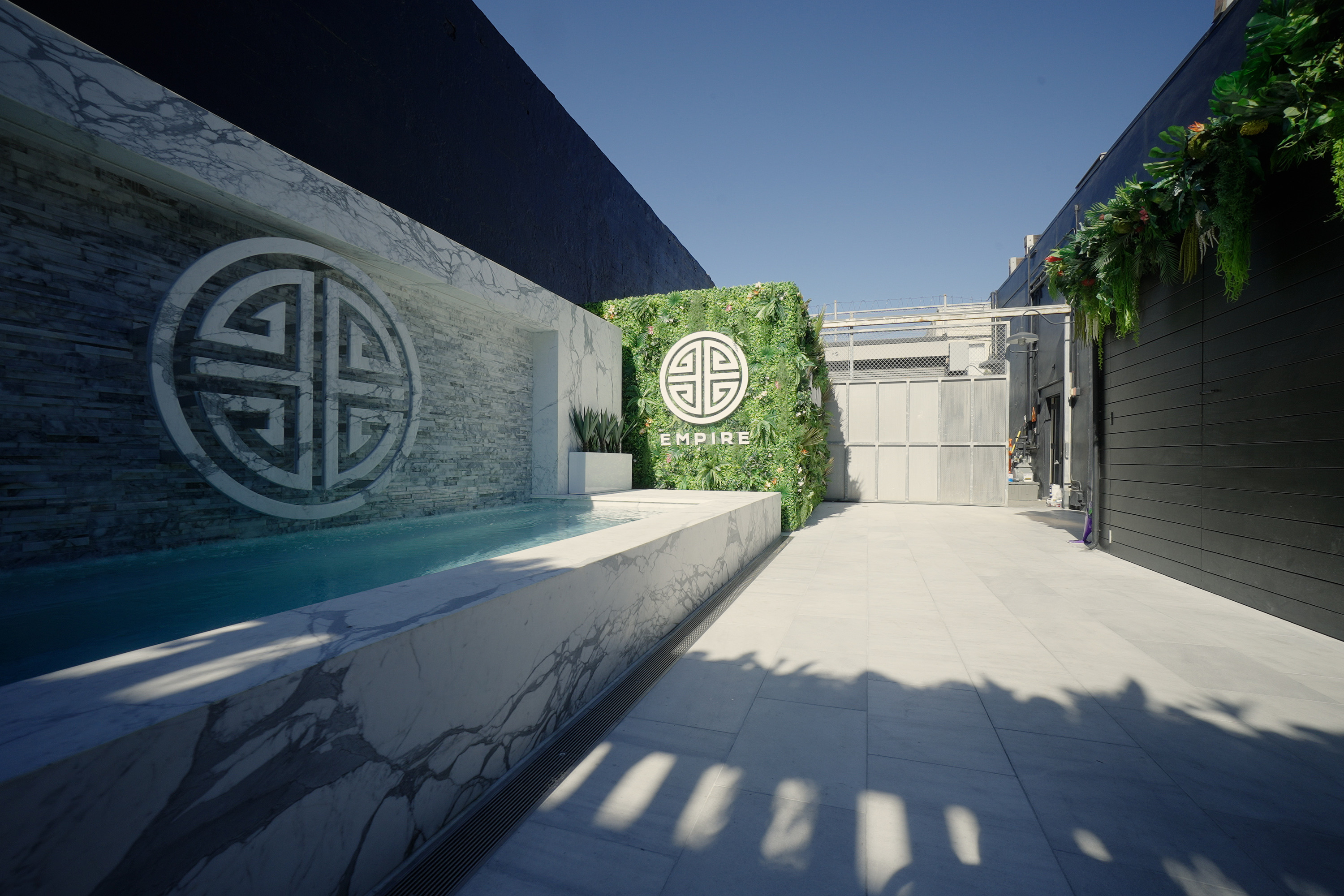 A sleek outdoor space with a small pool, marble walls, and a vertical garden. Two circular logos are displayed prominently on the wall.