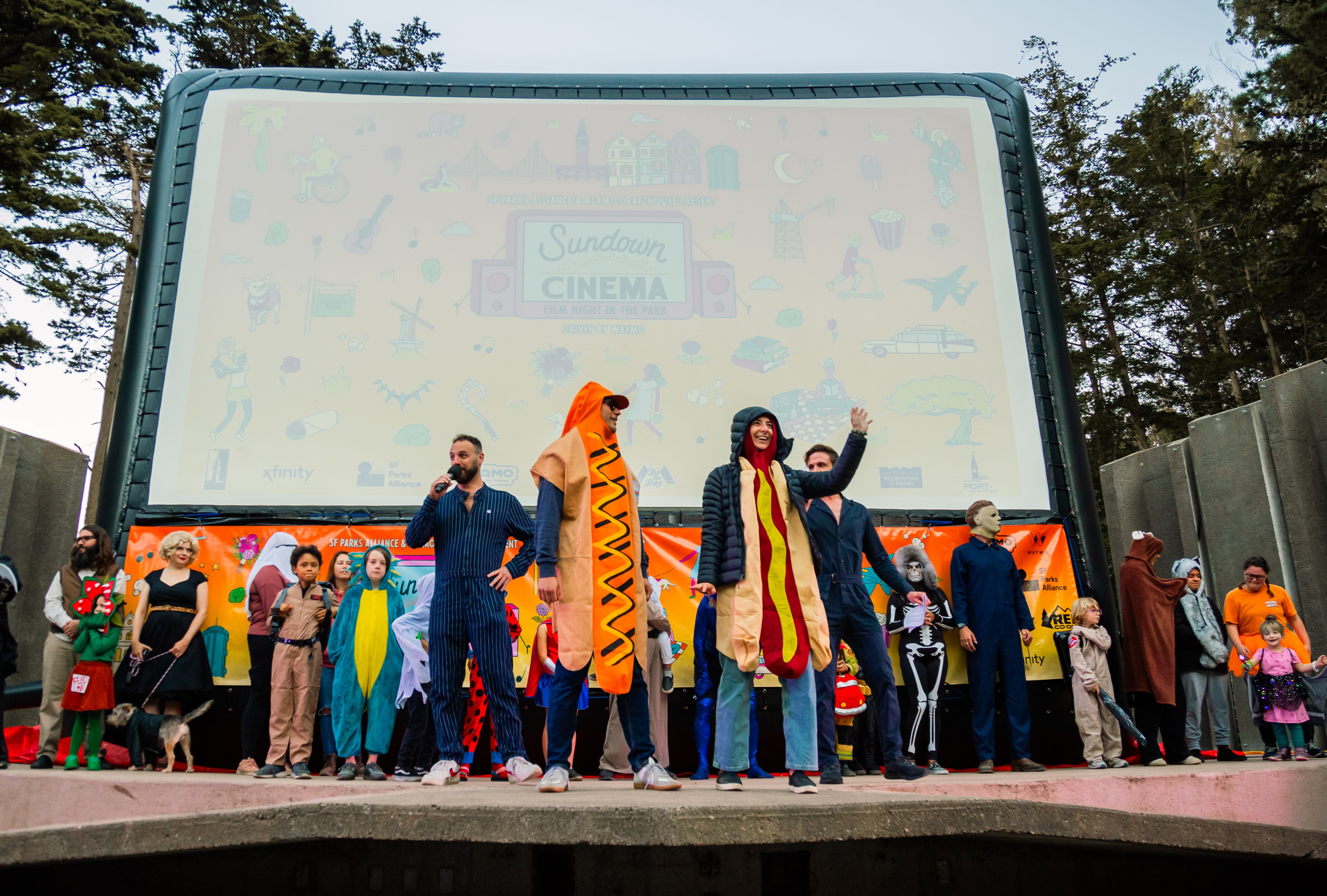 A group of people in colorful and humorous costumes stand in front of a large outdoor movie screen with the words &quot;Sundown Cinema&quot; displayed.