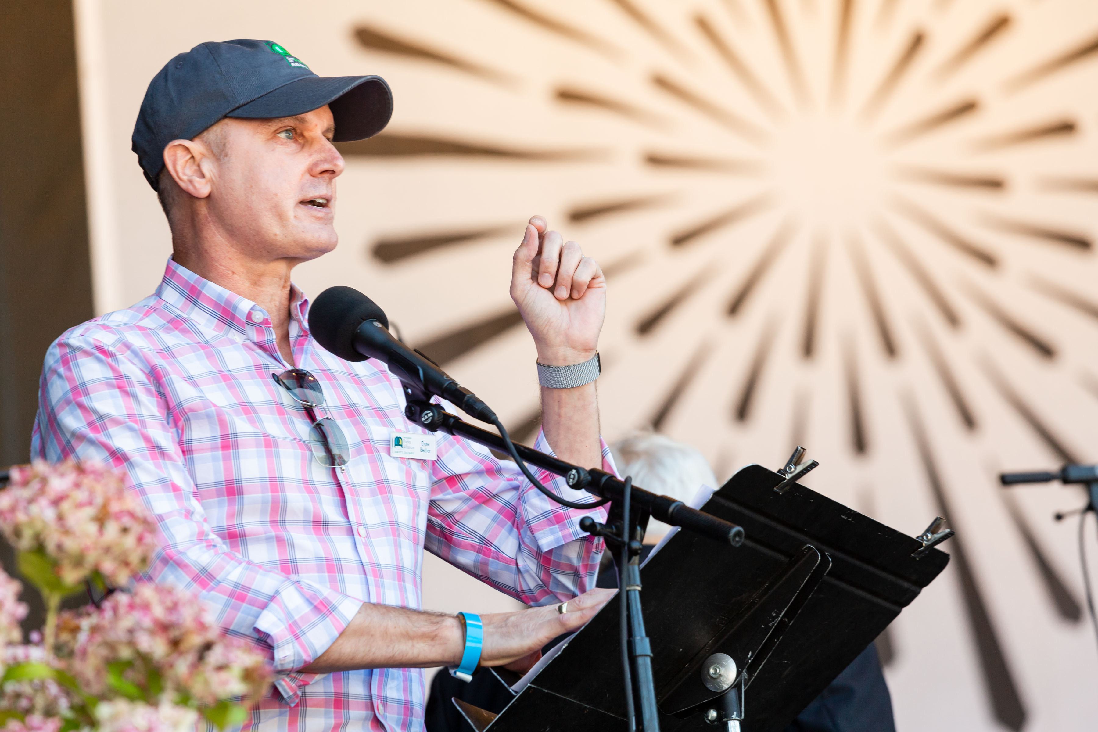 A person in a plaid shirt and cap speaks at a microphone on a stage, gesturing with one hand. Bright rays are depicted on a background wall behind him.