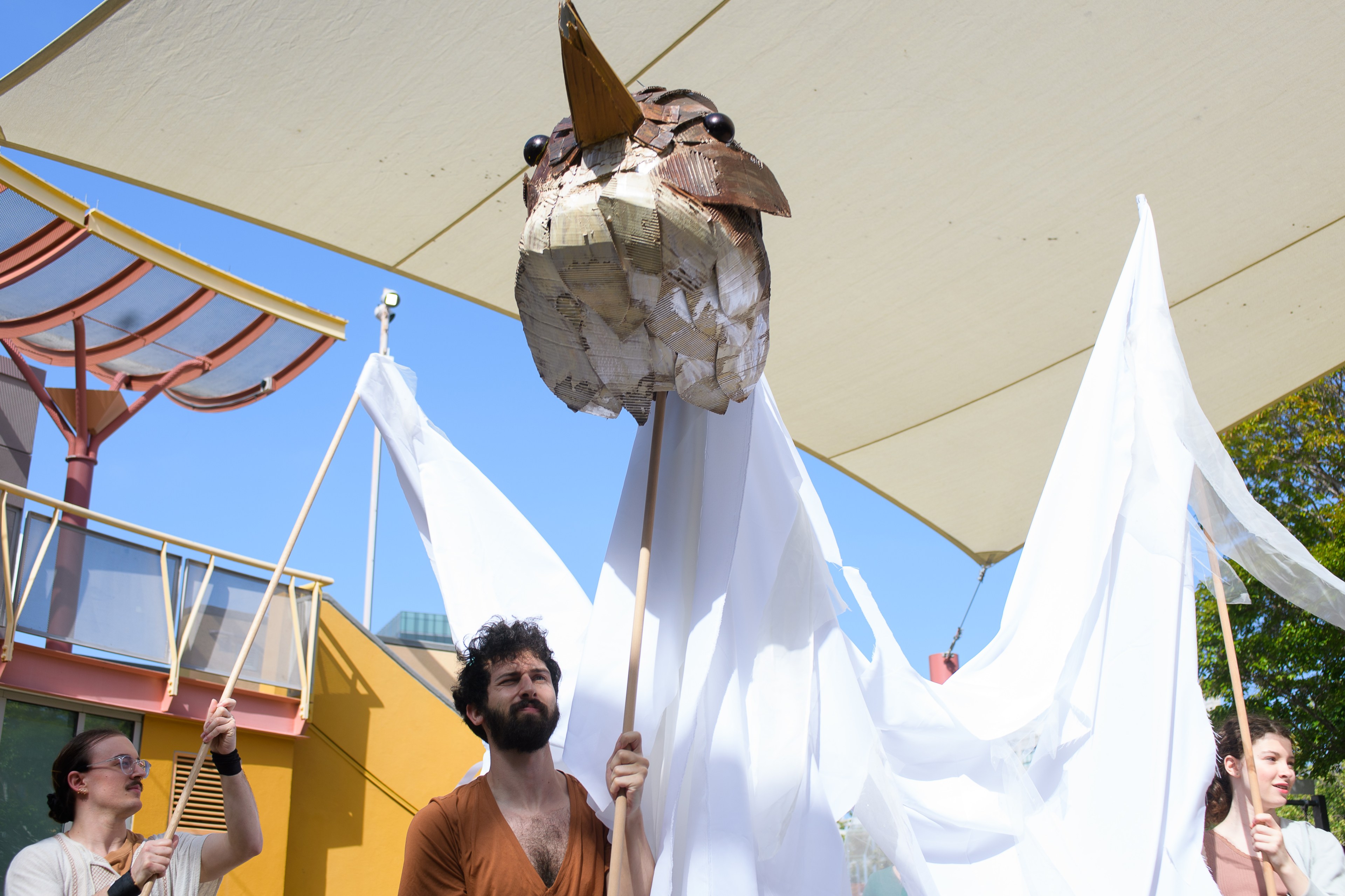 Three people hold up a large, paper-mache bird head and white fabric wings, creating the appearance of a giant bird. They stand outside under a canopy.