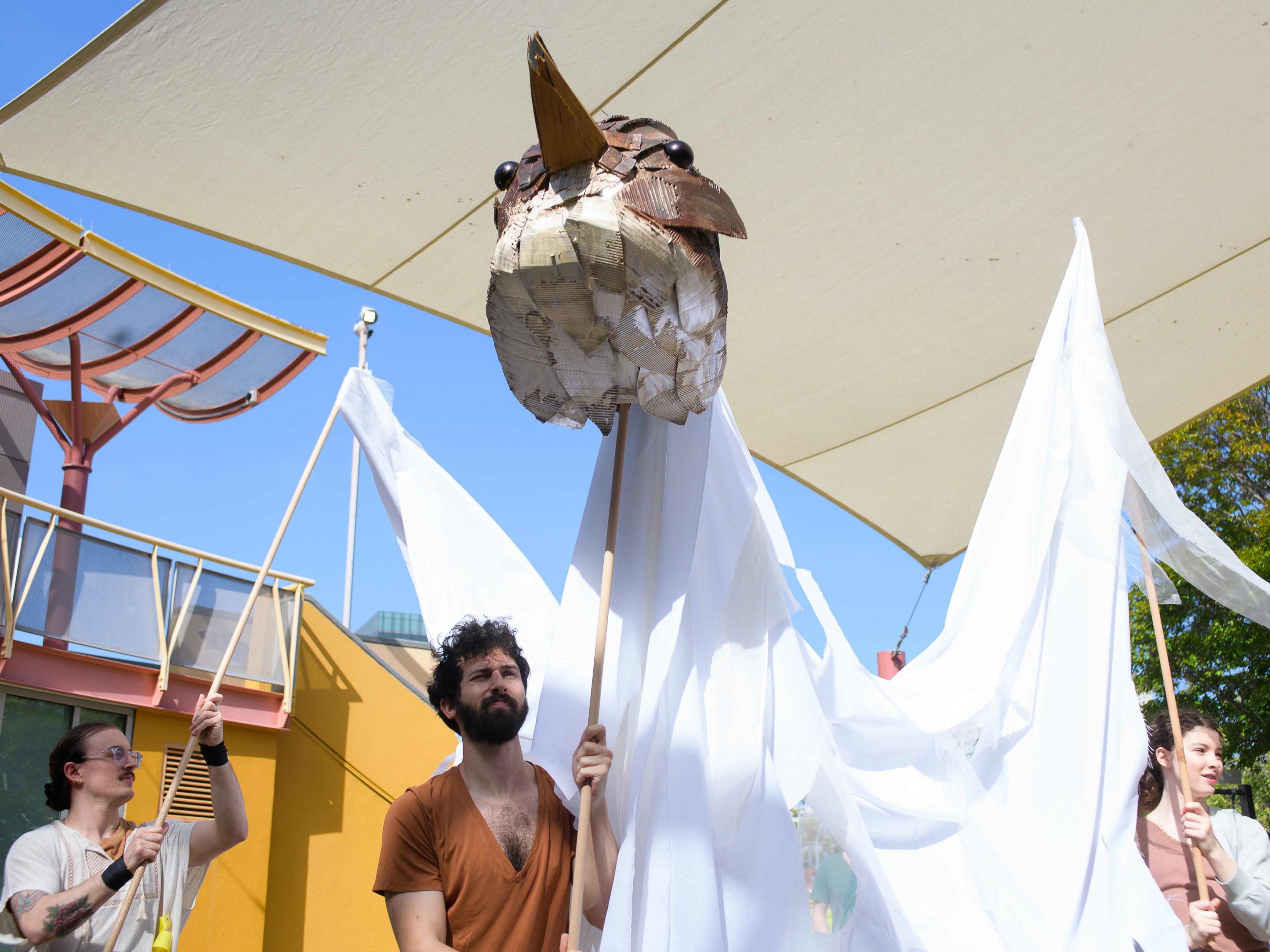 Three people hold up a large, paper-mache bird head and white fabric wings, creating the appearance of a giant bird. They stand outside under a canopy.