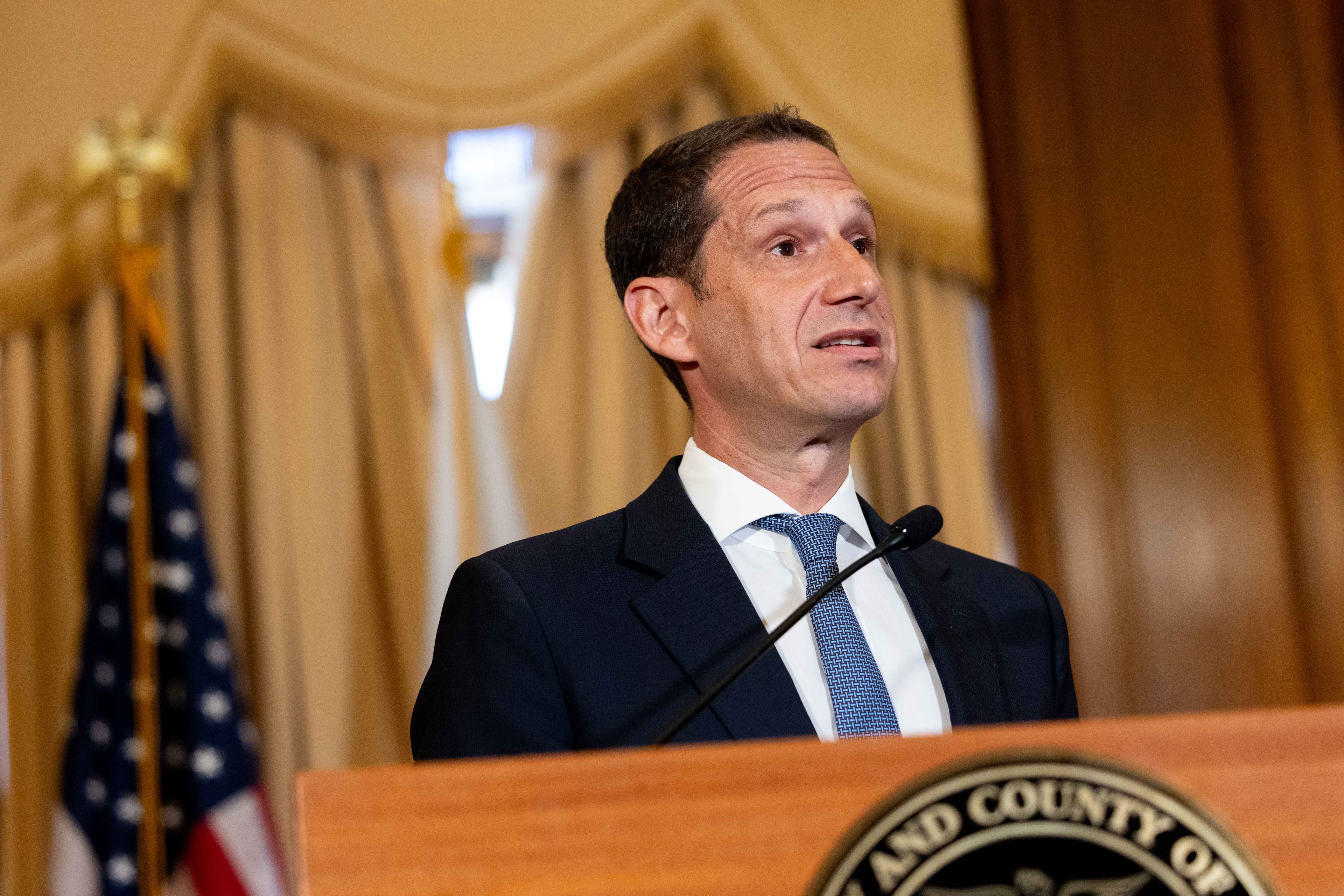 A man in a suit and tie speaks at a podium with a microphone. An American flag is visible in the background, along with curtains and a wood-paneled wall.