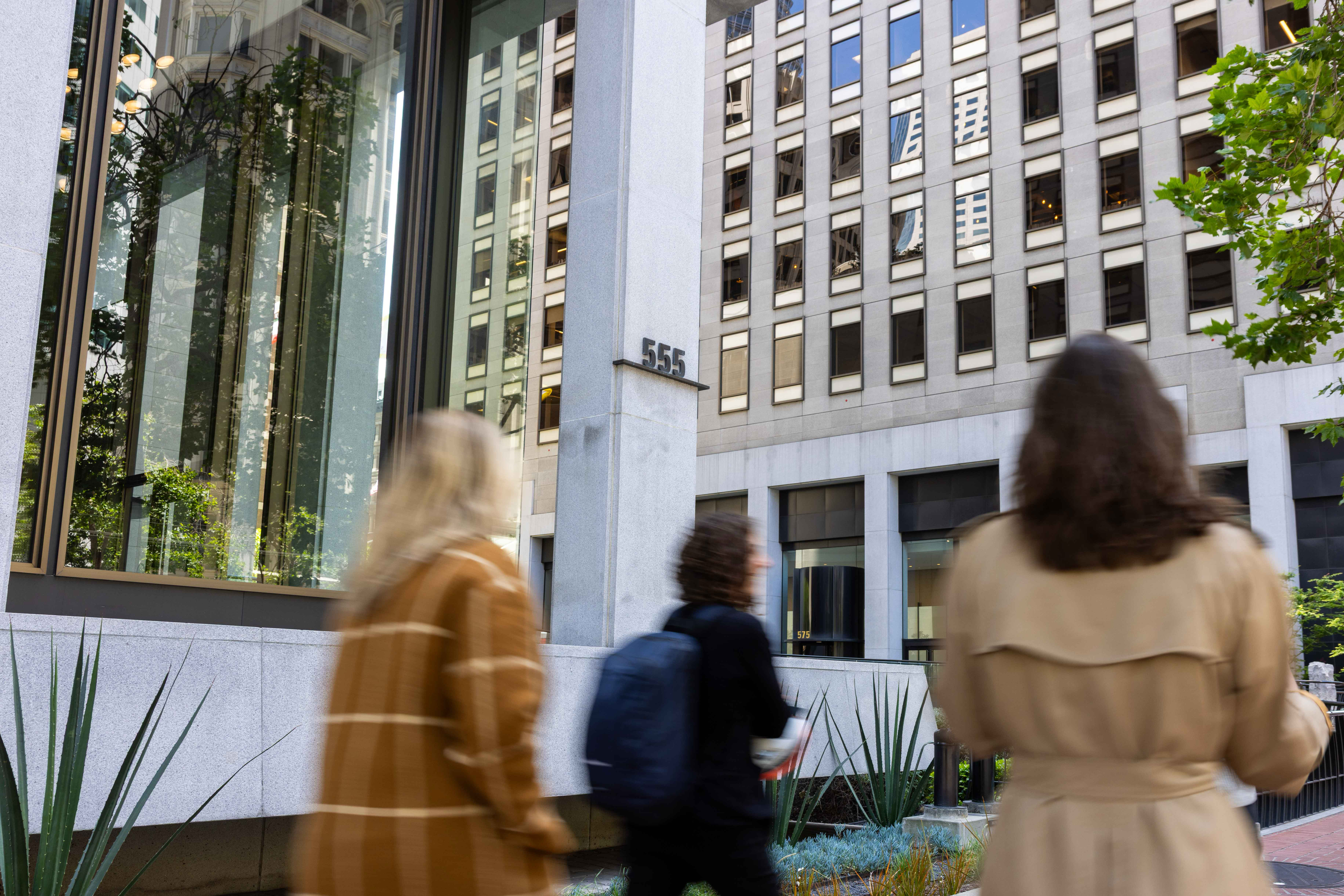 Three people in coats walk past a building with a glass facade, reflecting windows and trees. The building number &quot;555&quot; is visible on a column.
