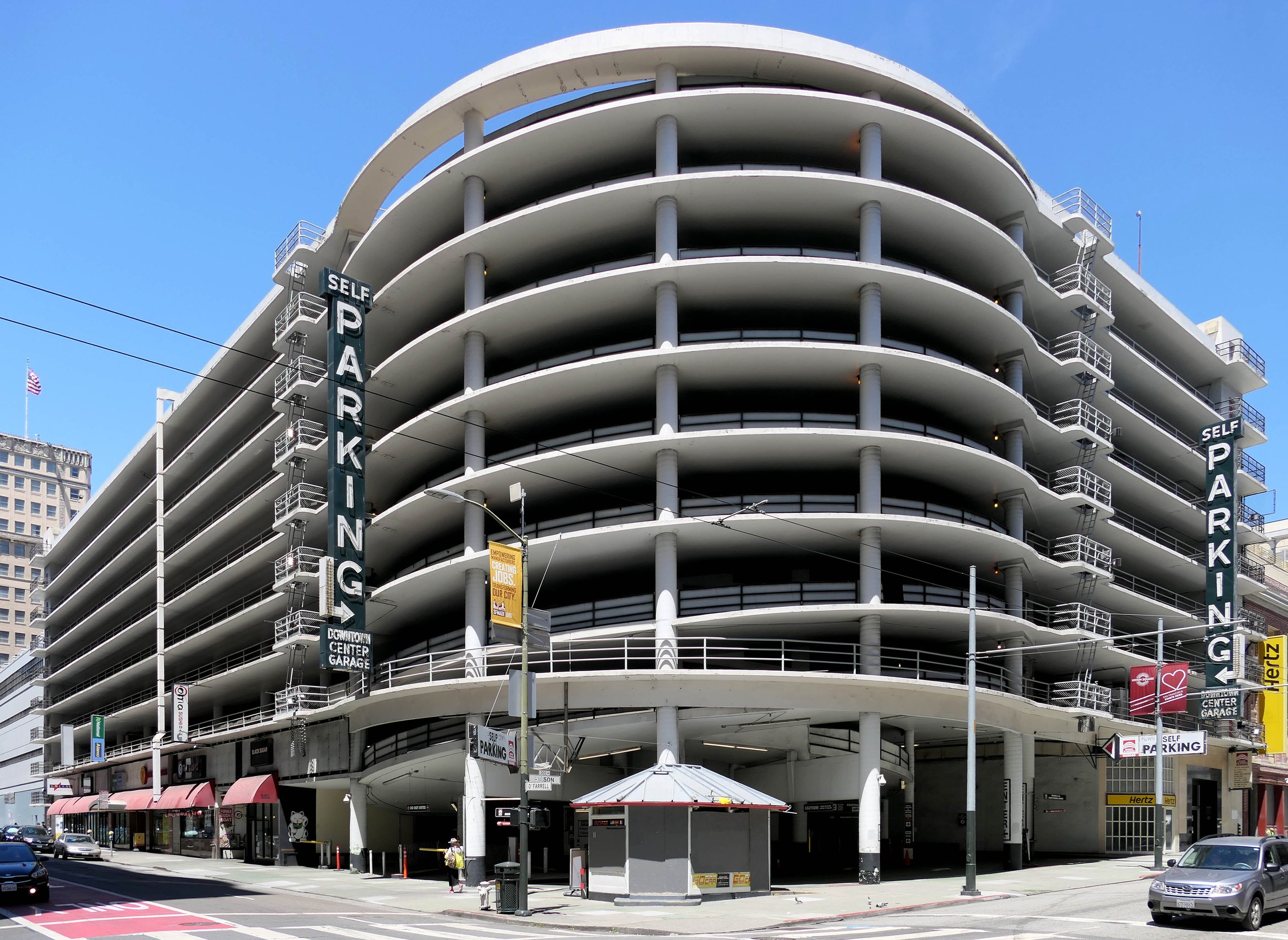 The image shows a multi-story, circular parking garage with signs saying &quot;Self Parking.&quot; It has open sides and storefronts at street level.