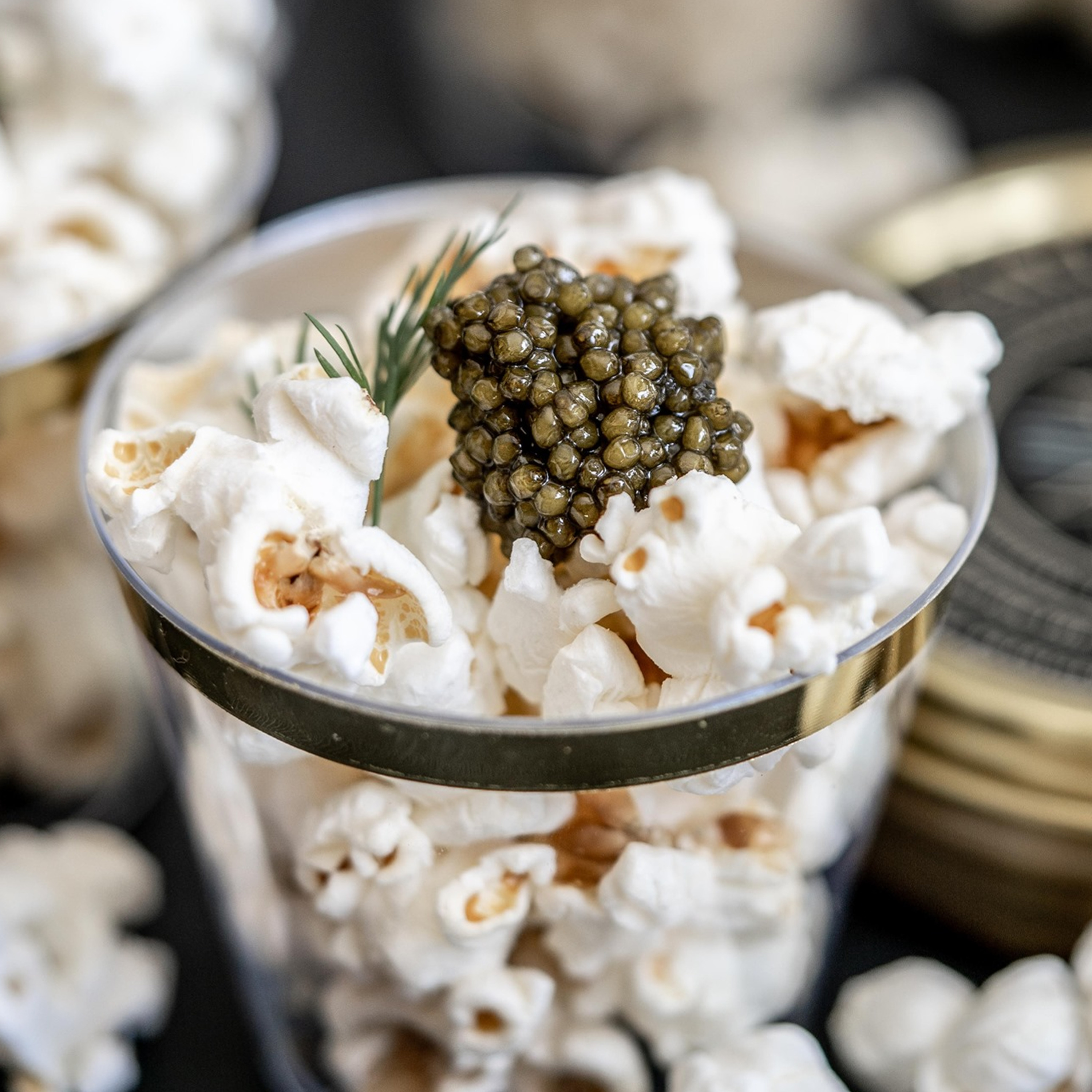 A clear cup is filled with popcorn, topped with a small mound of caviar and garnished with a sprig of dill.