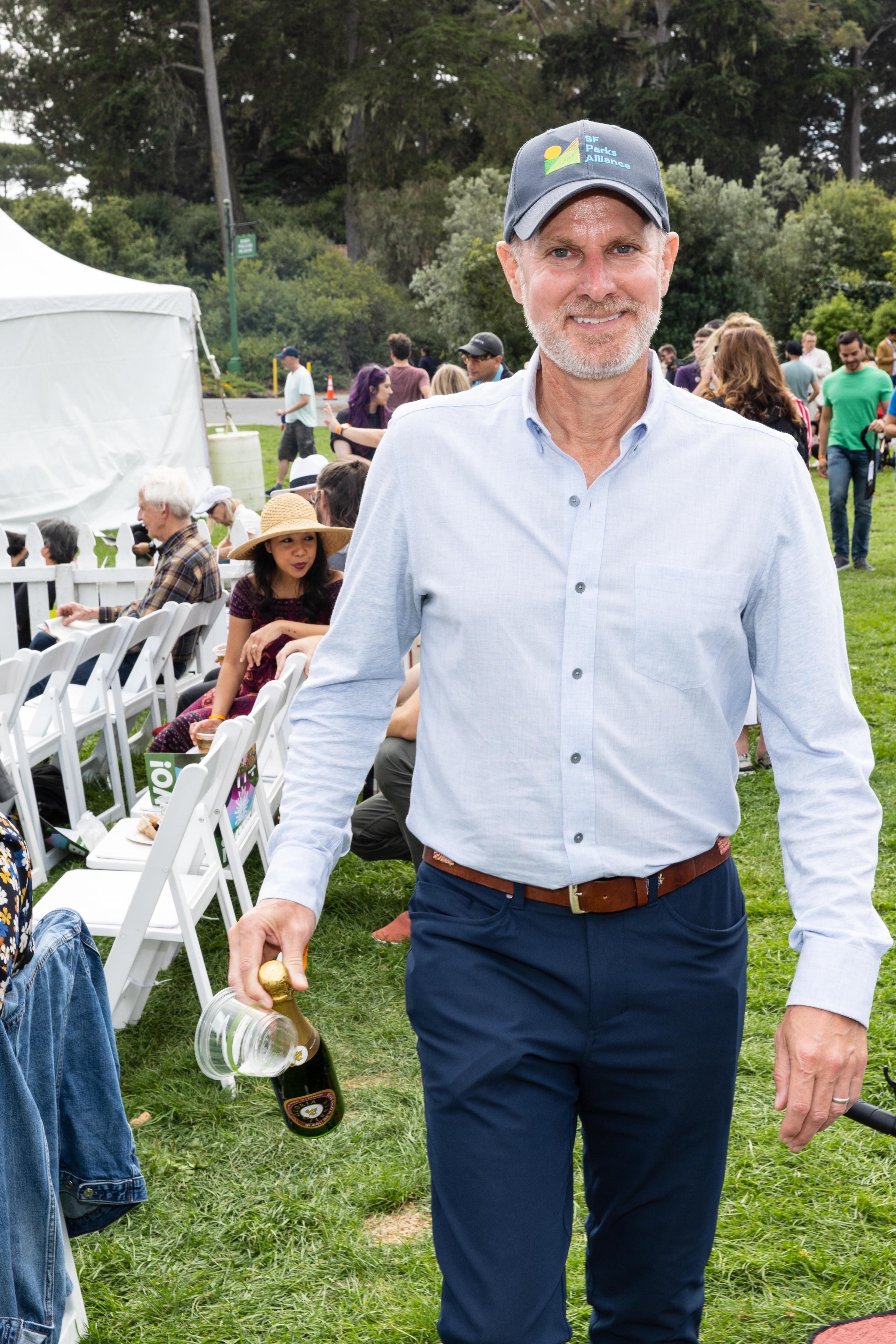 A man in a blue shirt and cap holds a bottle and cups at an outdoor event with people seated and standing on grass, surrounded by trees and a tent.
