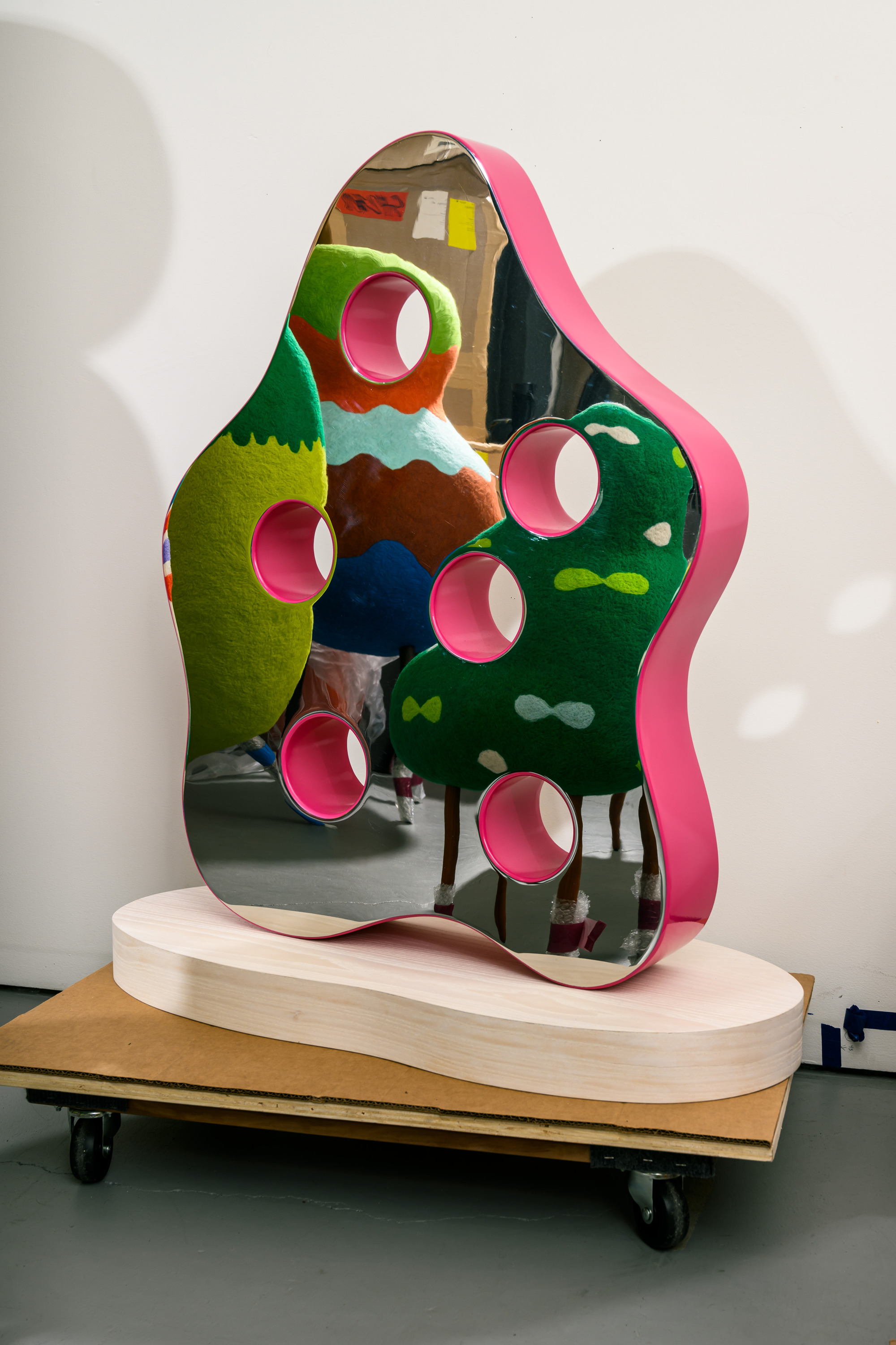 The image shows a pink, abstract sculpture with mirrored surfaces and circular cutouts, reflecting vibrant green and orange shapes, placed on a wooden base.
