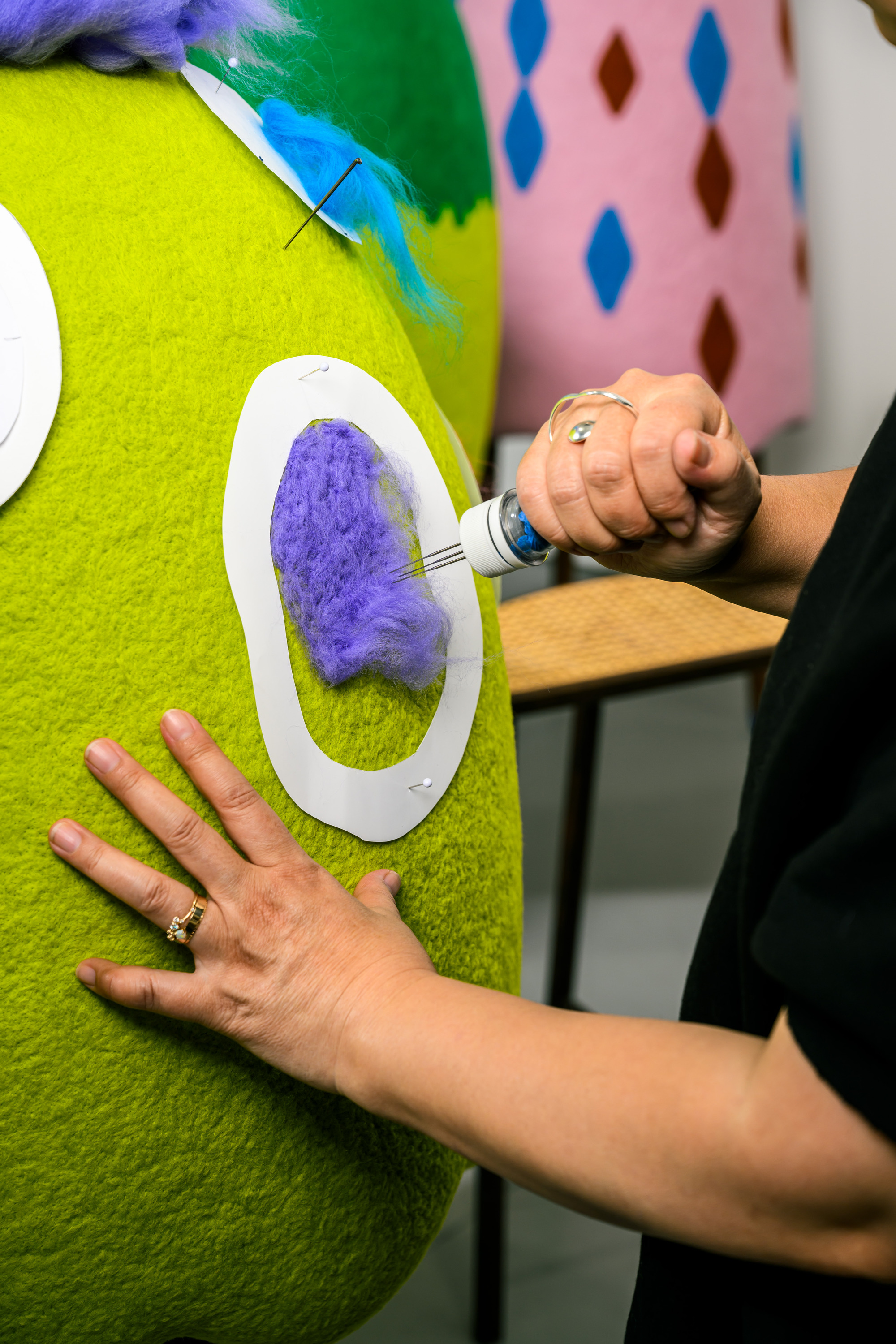 A person needle-felts purple wool onto a green felt surface, using a stencil to guide the design. There's a felt object in the background with colorful patterns.