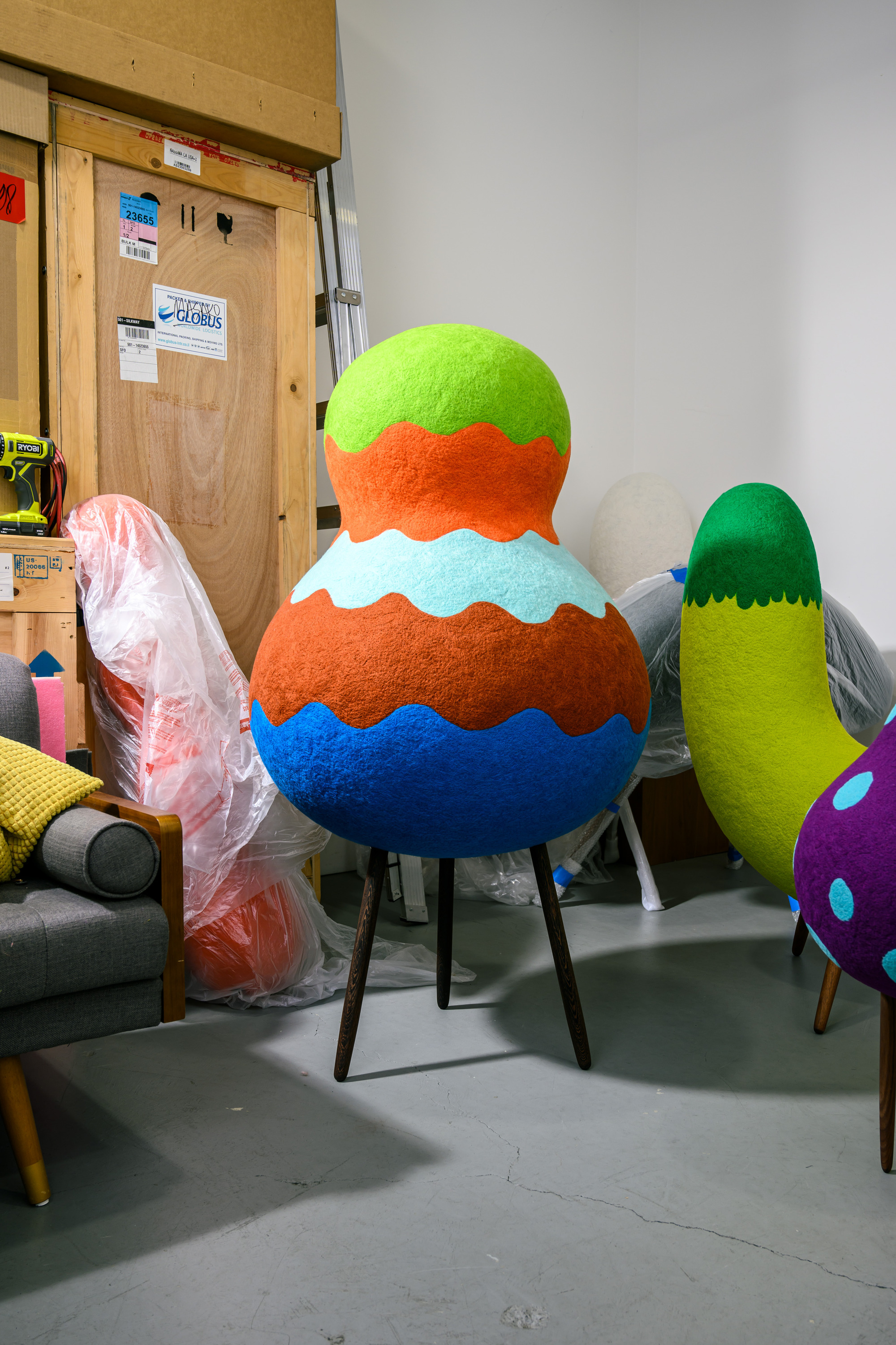 The image shows colorful, abstract sculptures with wavy patterns on legs in a cluttered room with boxes and a gray chair nearby.