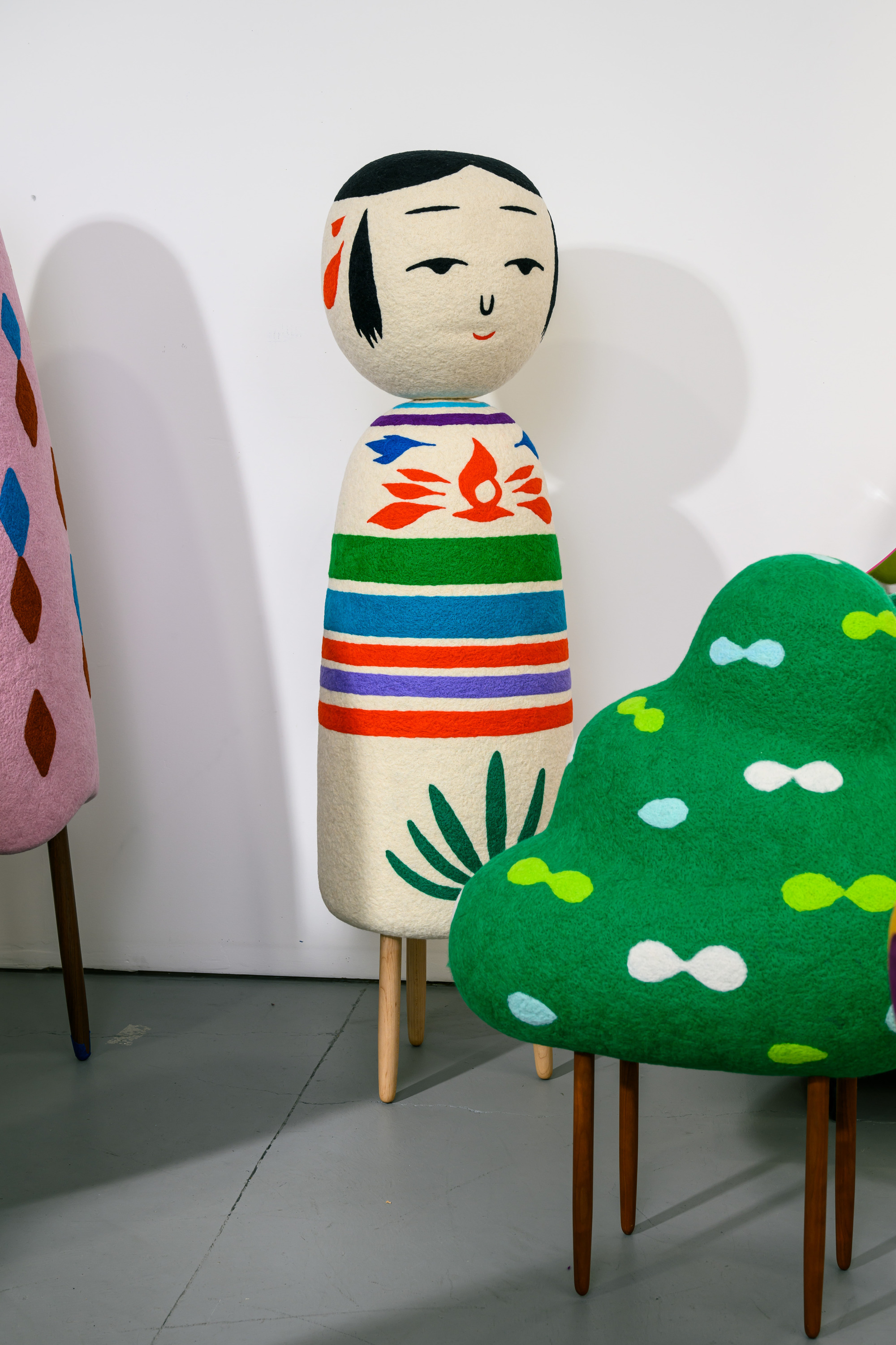 The image shows a colorful, abstract sculpture shaped like a doll with a painted face. It has a vibrant, striped body with decorative patterns.