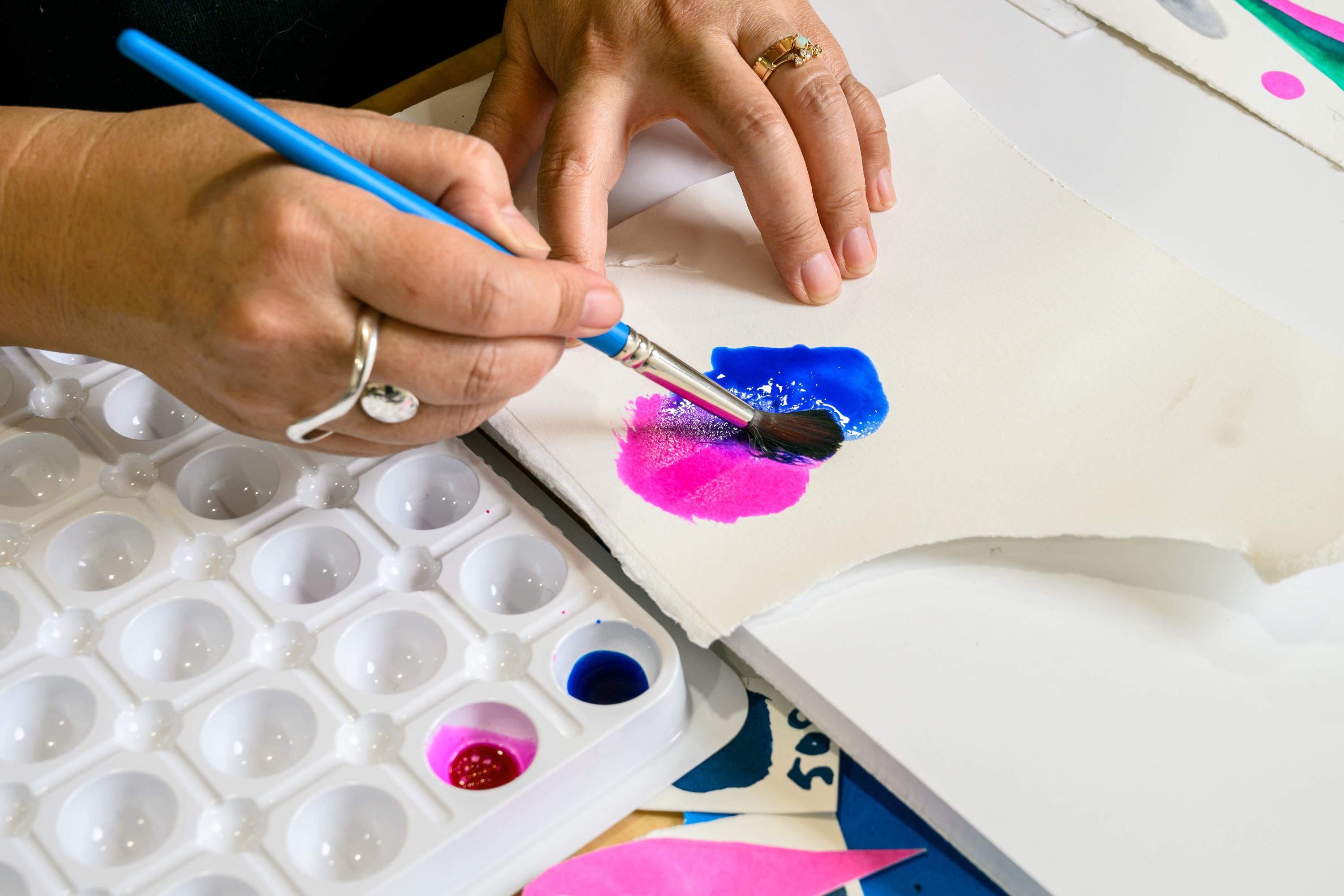 A person is painting with a blue brush on paper, blending blue and pink colors. A palette with small paint wells is visible nearby.