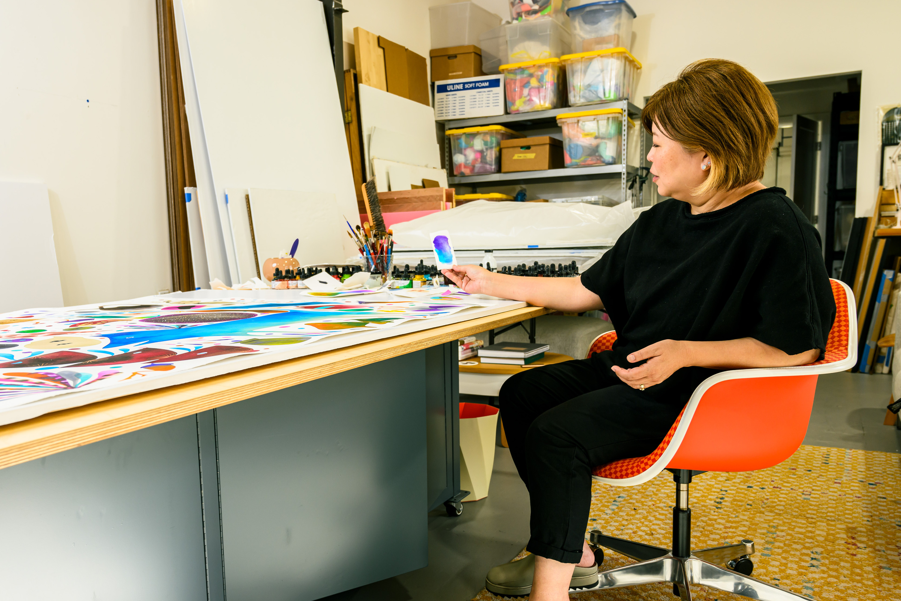 A woman sits on an orange chair in an art studio, examining a colorful painting on a table. The room is filled with art supplies and storage boxes.