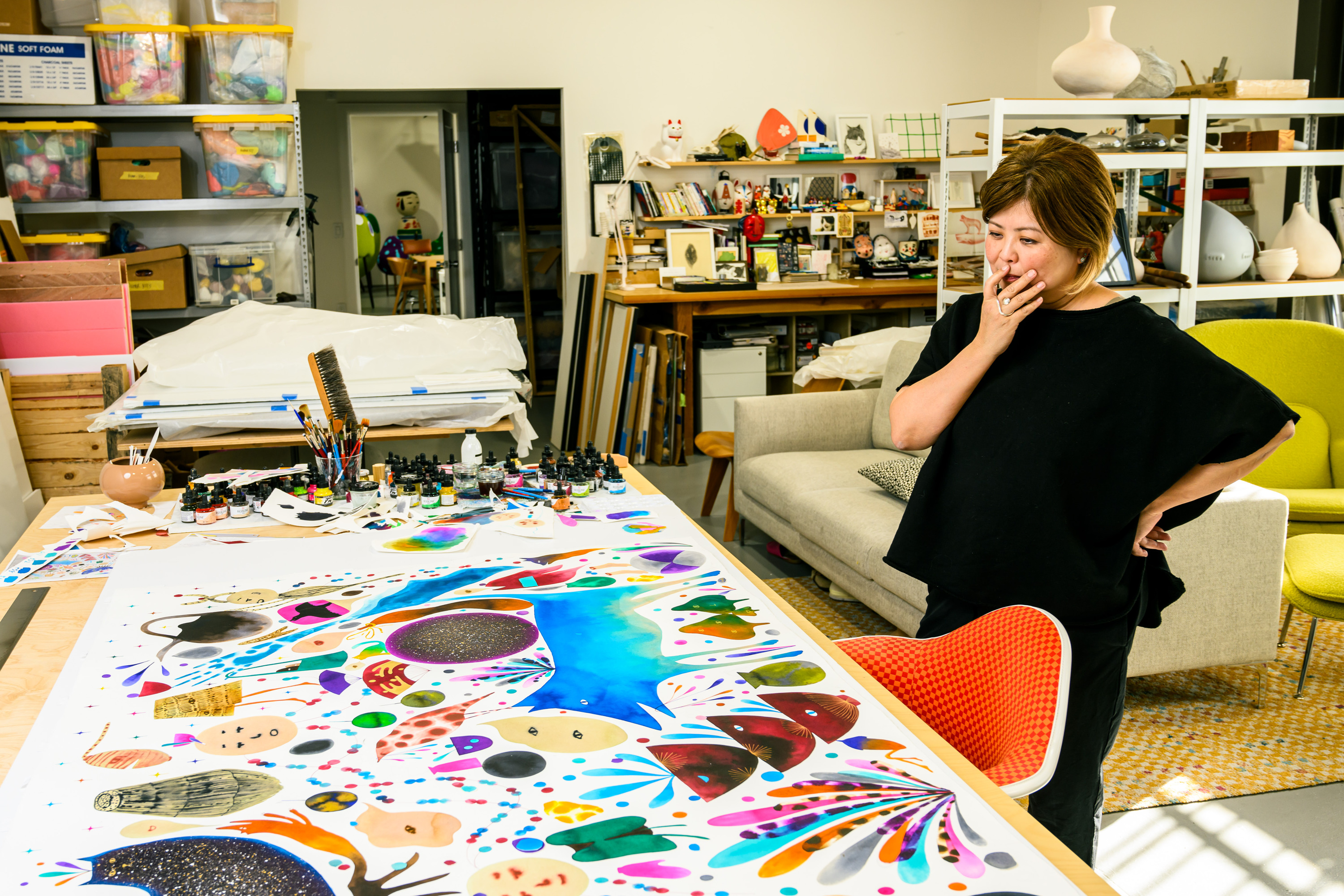 A woman stands thoughtfully in a vibrant art studio, surrounded by colorful abstract artwork and various art supplies on a cluttered table.
