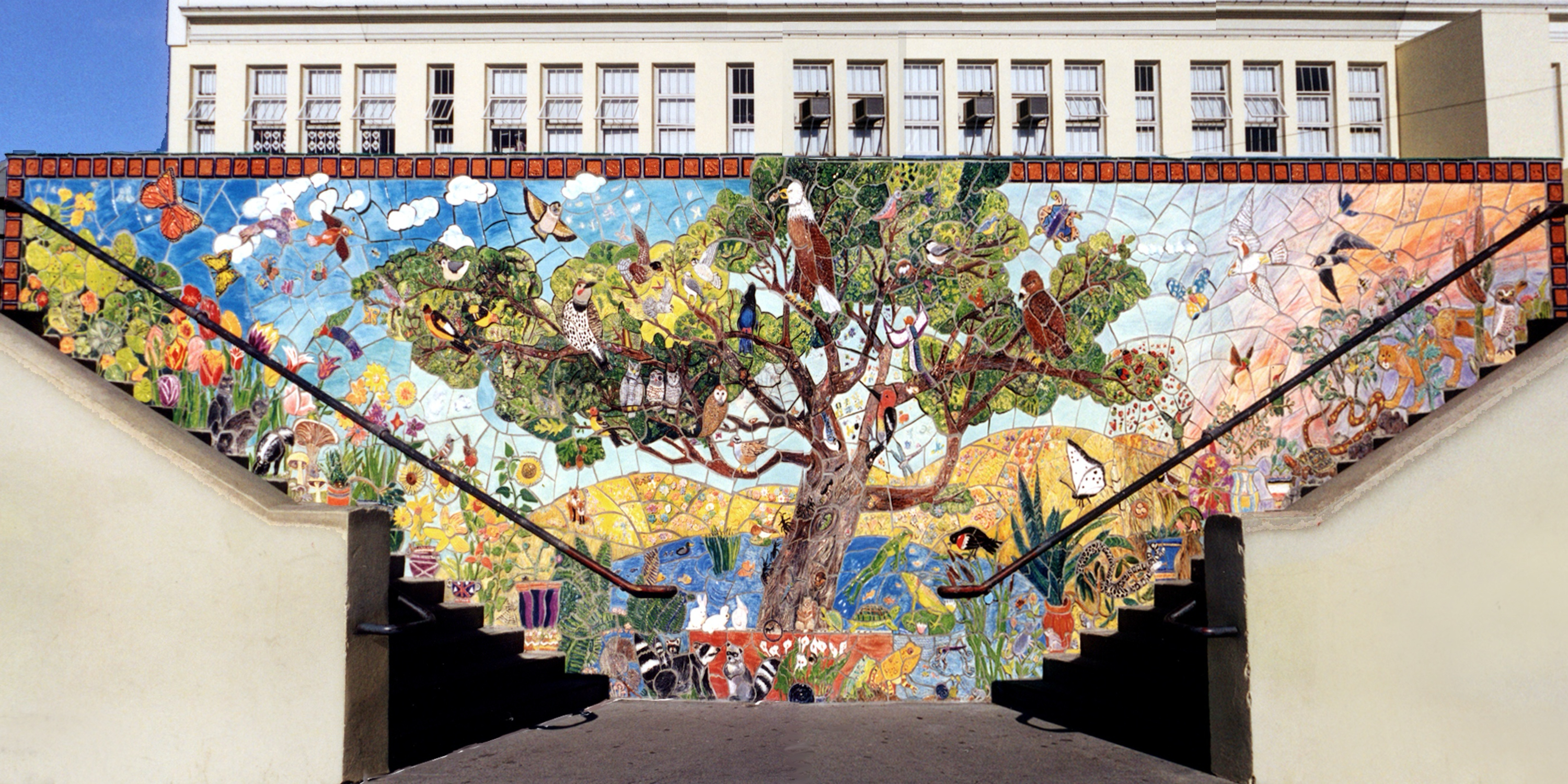 The image shows a vibrant mosaic mural featuring a large tree filled with various birds and surrounded by colorful plants, flowers, and animals against a blue sky.
