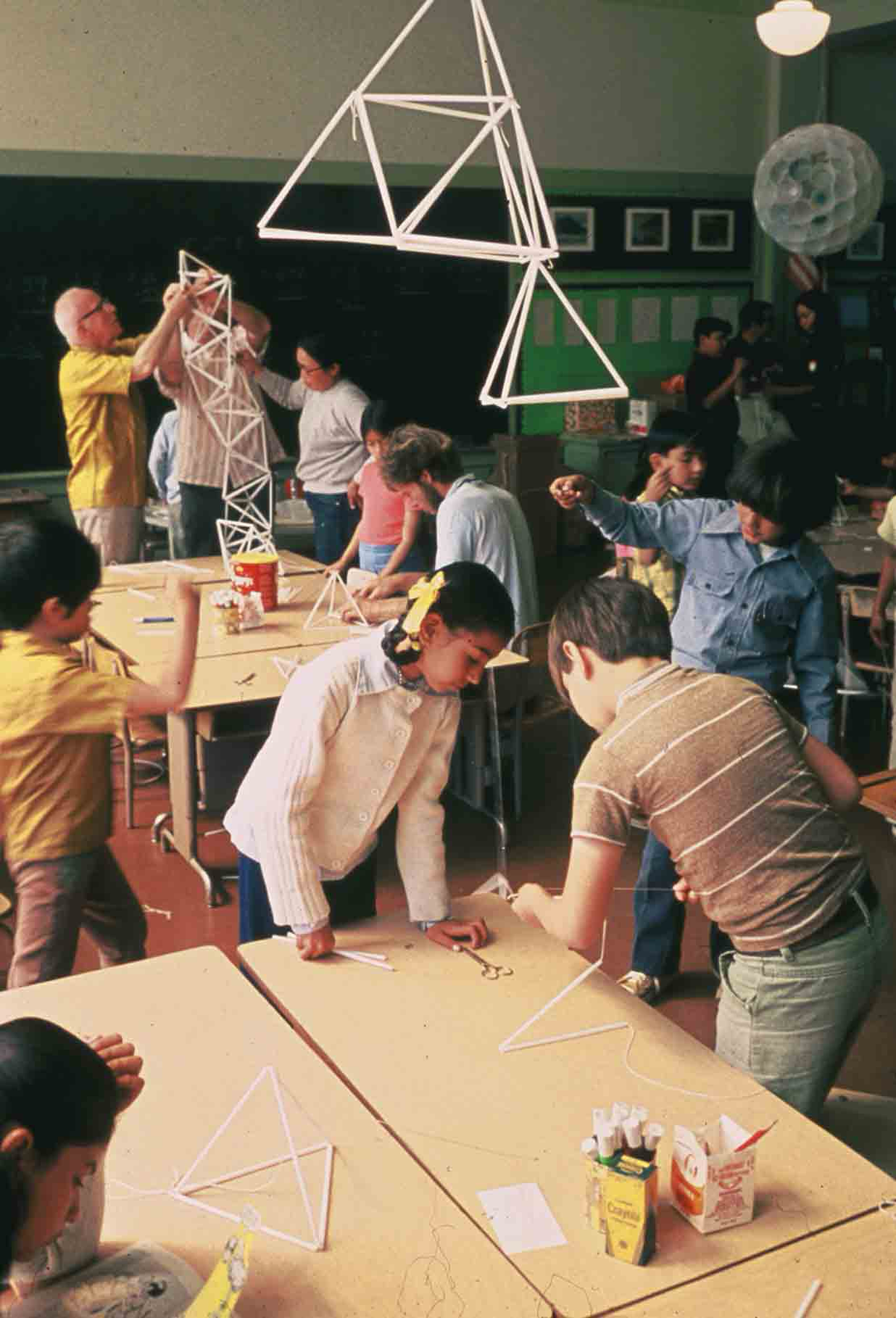 Children and adults are working on geometric structures made from straws in a classroom. Tables are scattered with materials, and models hang from the ceiling.
