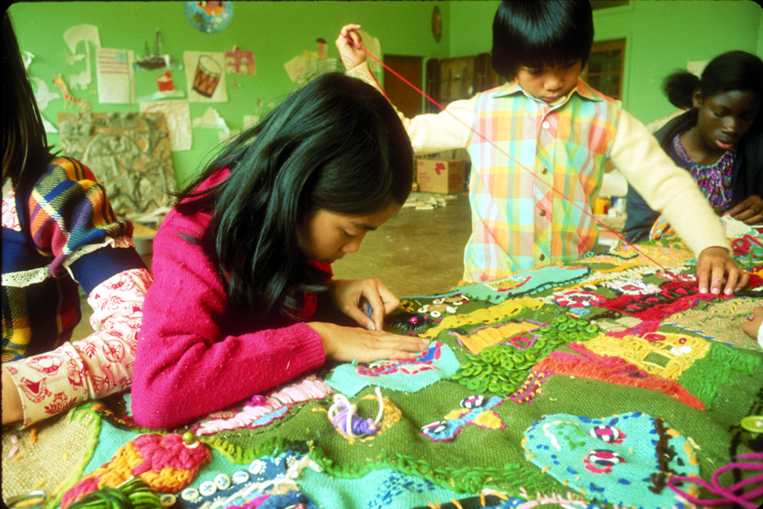 Children are focused on a colorful, detailed textile project, featuring buttons and yarn. The background displays drawings on a green wall.