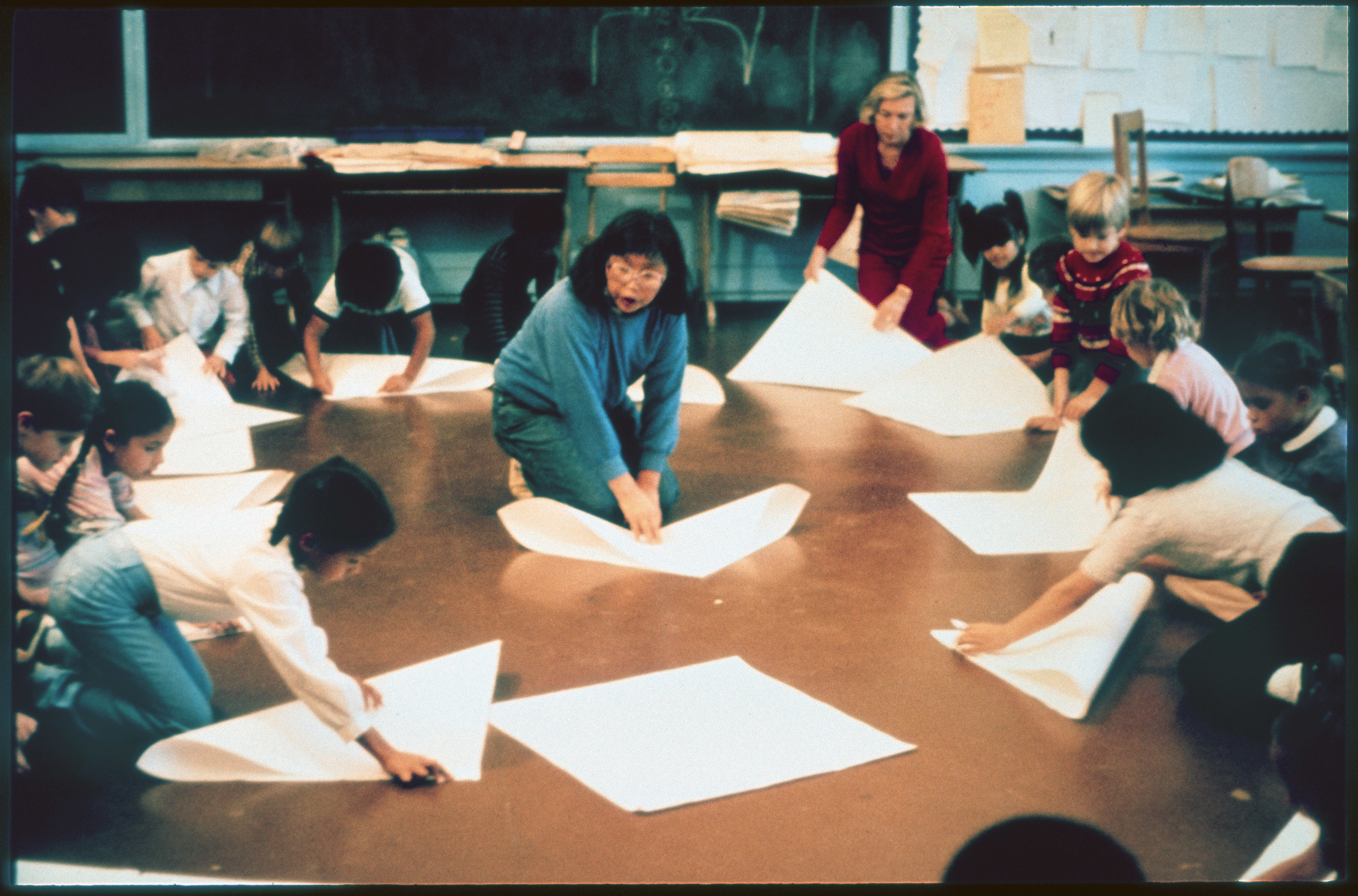 A group of children and two adults are on the floor, folding large sheets of paper in a classroom setting. They appear engaged and focused on the activity.