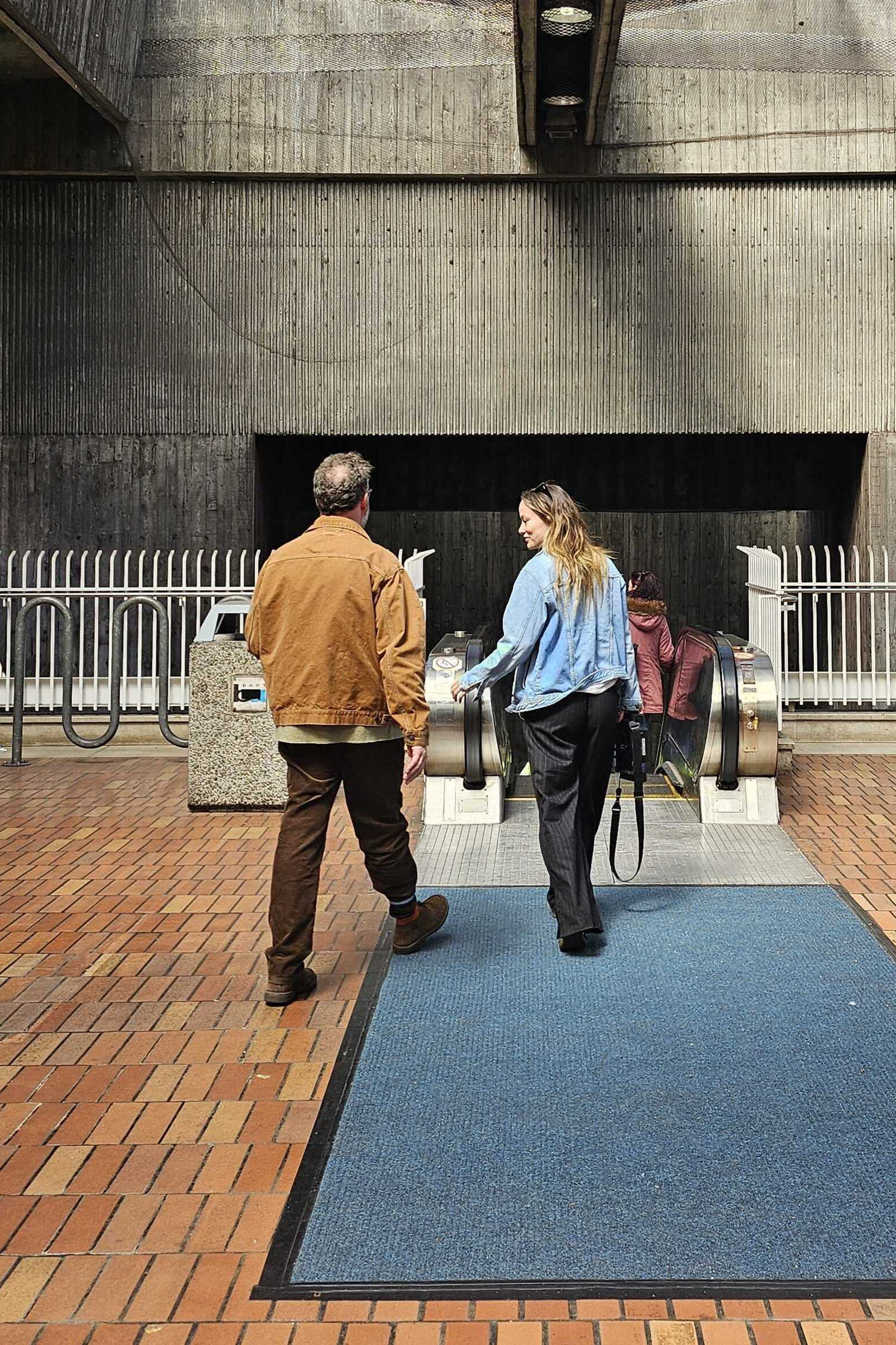A man and woman approach an outdoor escalator in a concrete setting. The man wears a brown jacket; the woman in a blue jacket carries a camera.