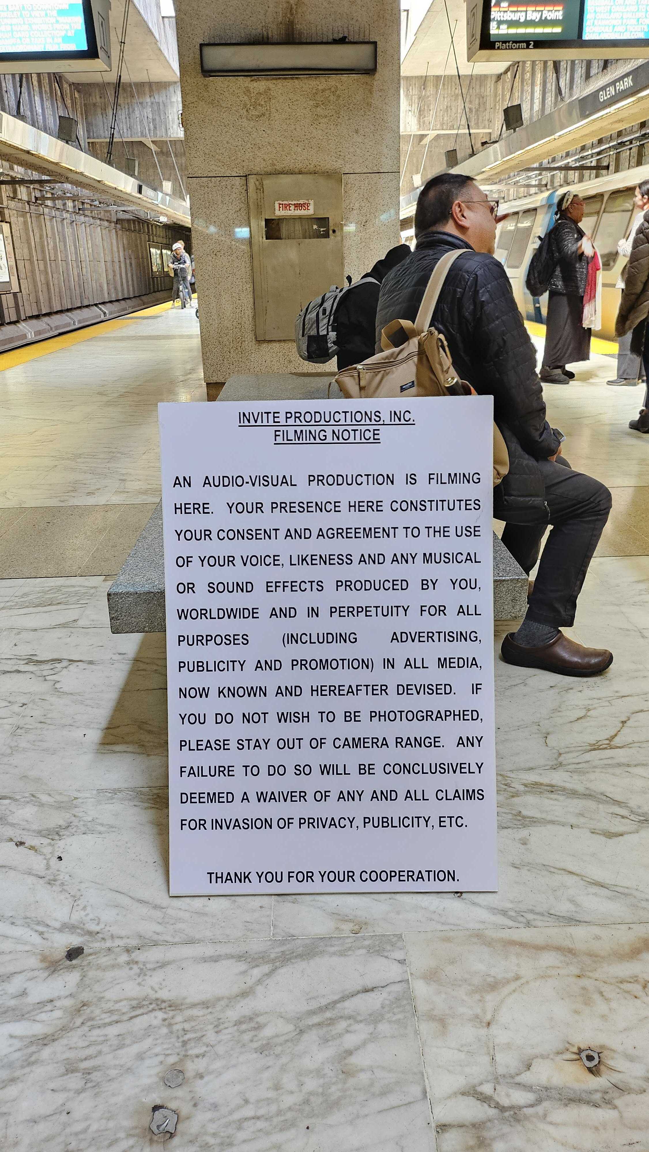 A subway station with a filming notice in the foreground. People are sitting and standing on the platform, with a train partially visible on the right.