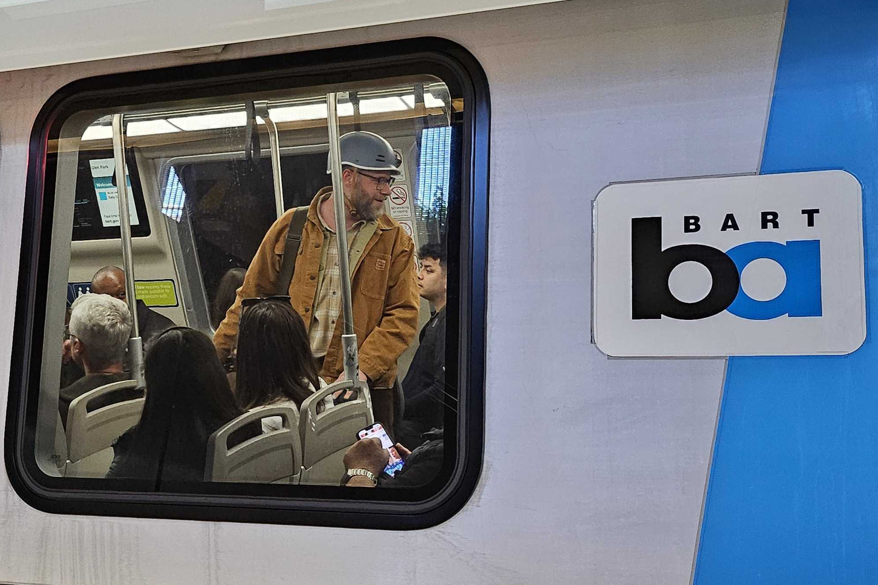 A group of people is seen sitting and standing inside a BART train. One man in a jacket and helmet stands, while others are seated, some using phones. The BART logo is visible outside.