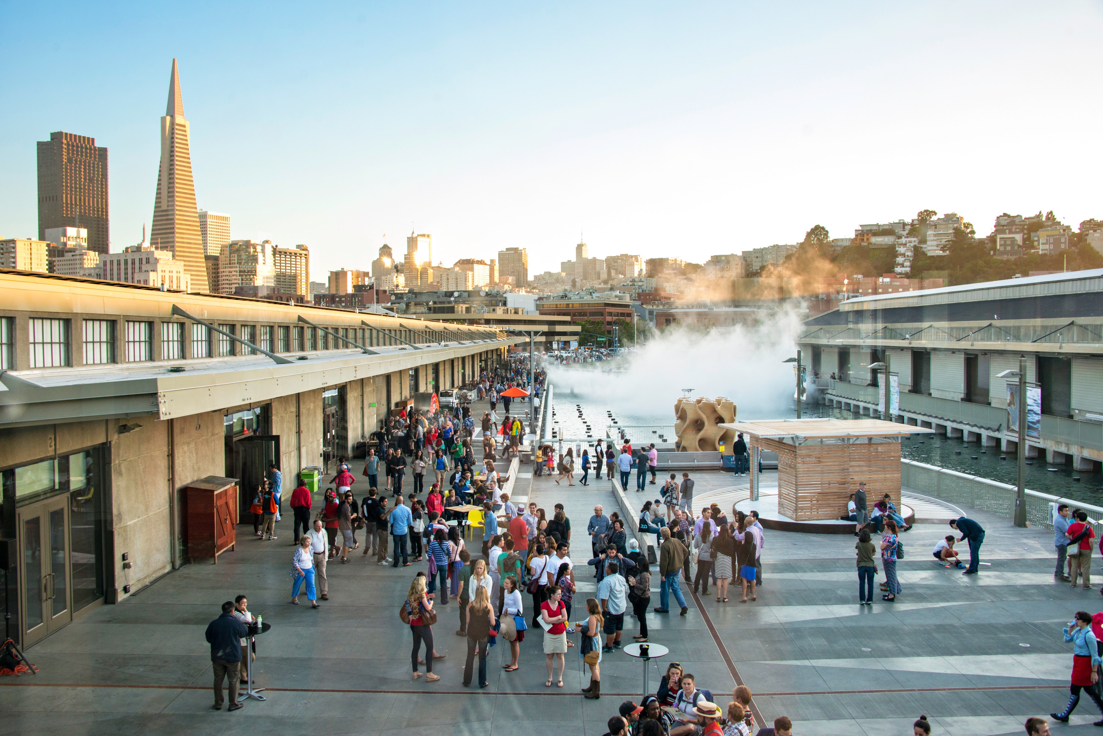 A bustling outdoor waterfront scene with many people gathered on a wide promenade. Skyscrapers are visible in the background, and mist rises near the docks.