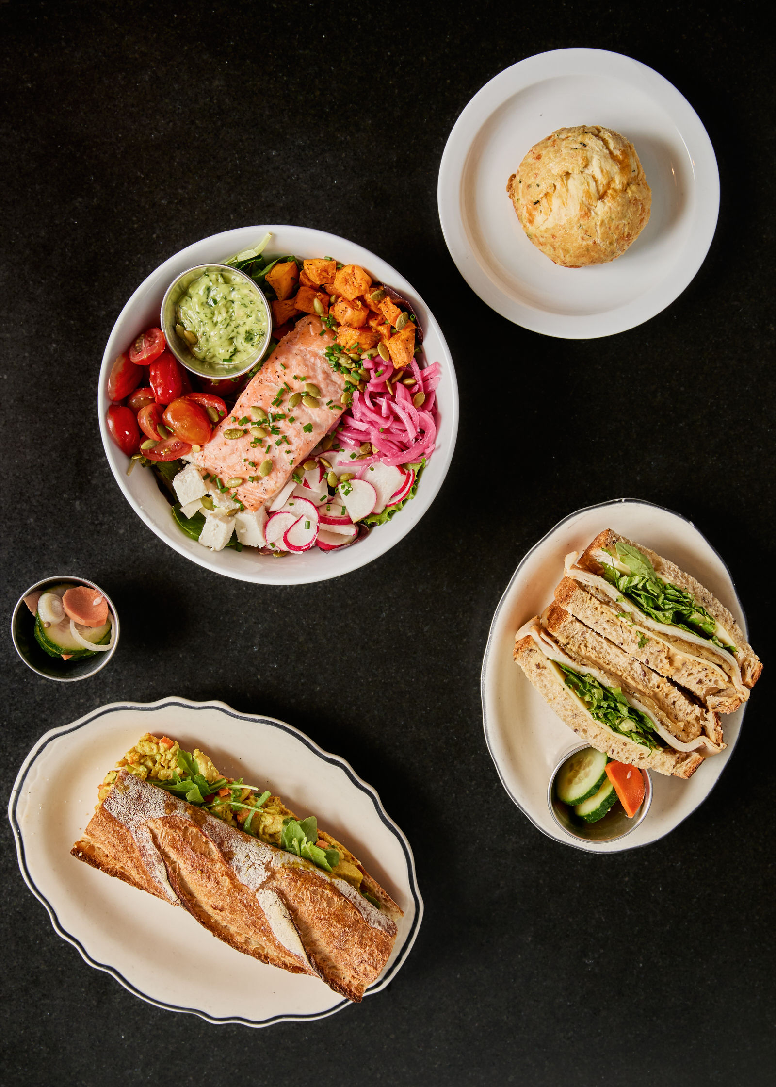 A salmon salad bowl with avocado, tomatoes, and radishes, a biscuit, a pickle cup, and two sandwiches on separate plates are arranged neatly on a dark surface.