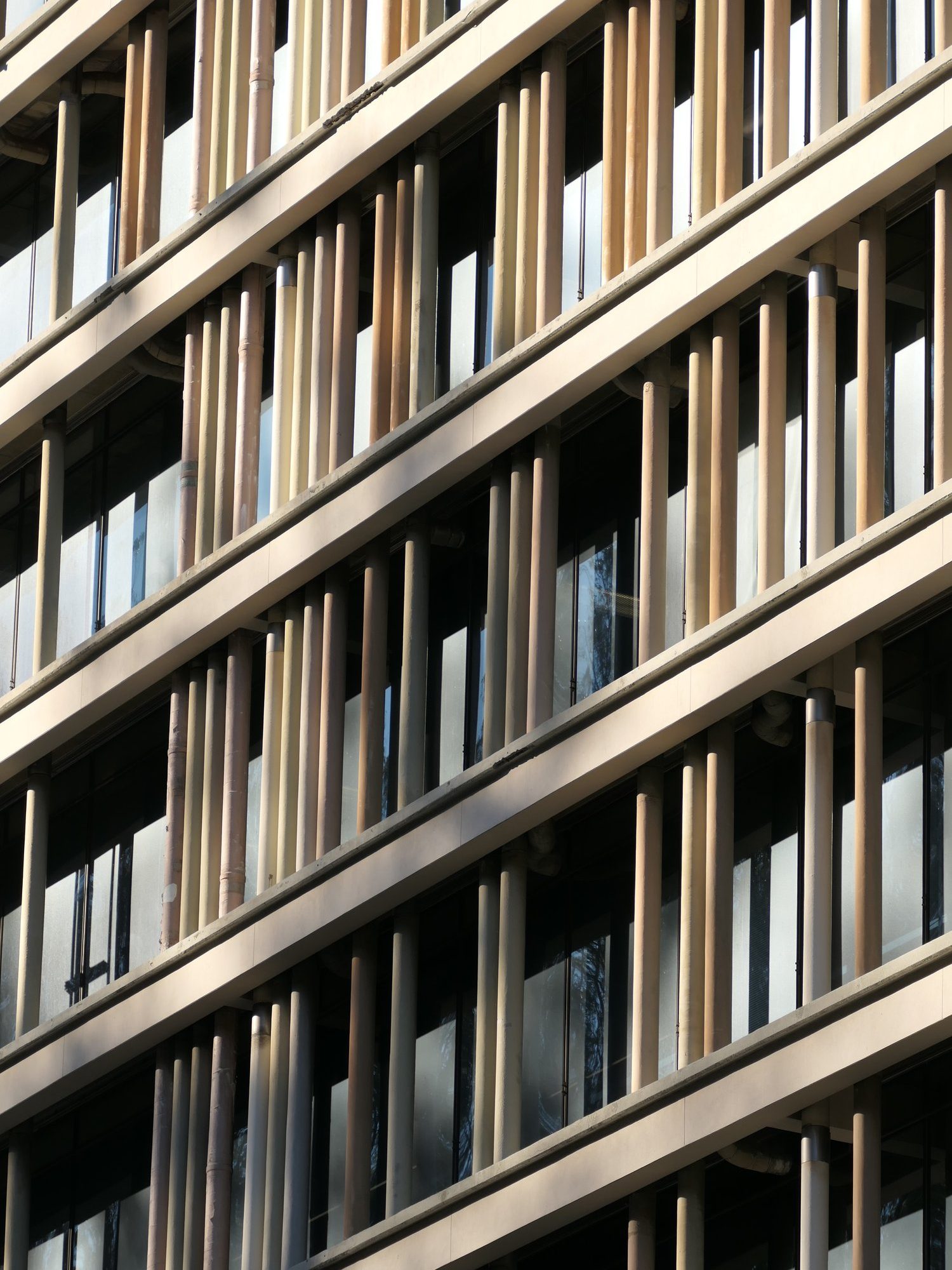 The image shows a building facade with vertical beige and brown slats aligned in a grid pattern, creating a geometric look against reflective glass windows.