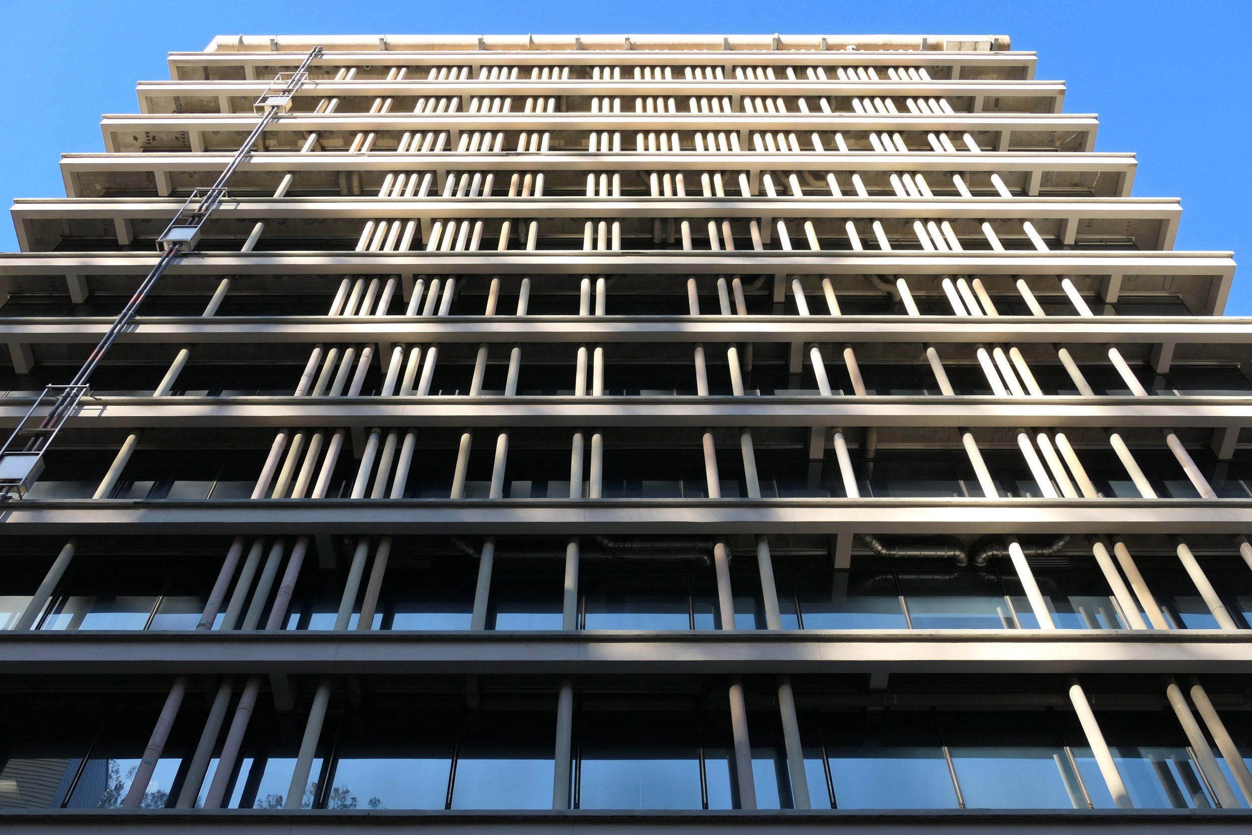 The image shows a tall, modern building with vertical white columns and multiple horizontal floors. The sky is clear and blue, and a narrow window washer's ladder is visible.