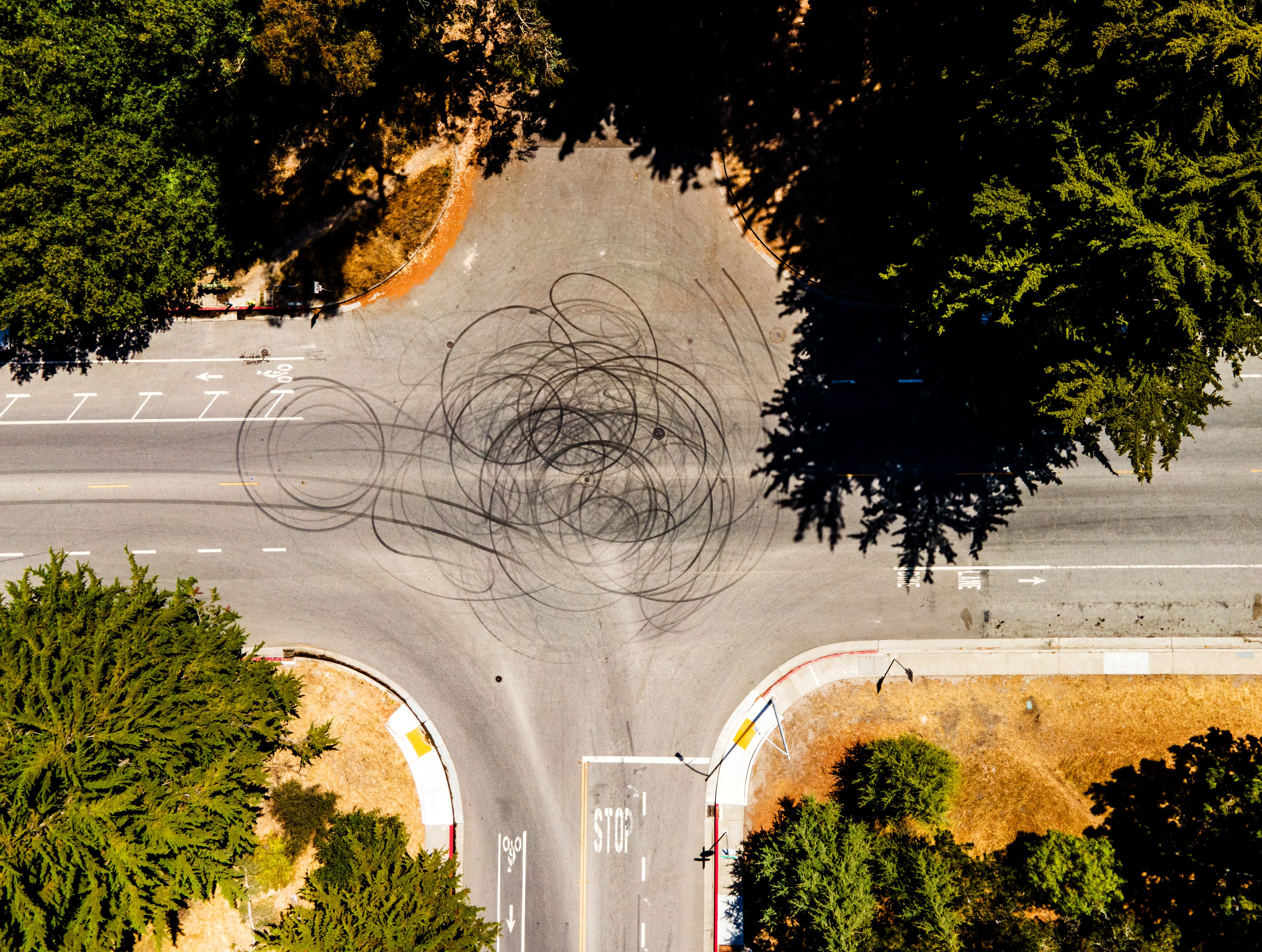 An aerial view of an intersection shows dark tire skid marks in circular patterns on the road. The area is surrounded by trees and some dry grass patches.