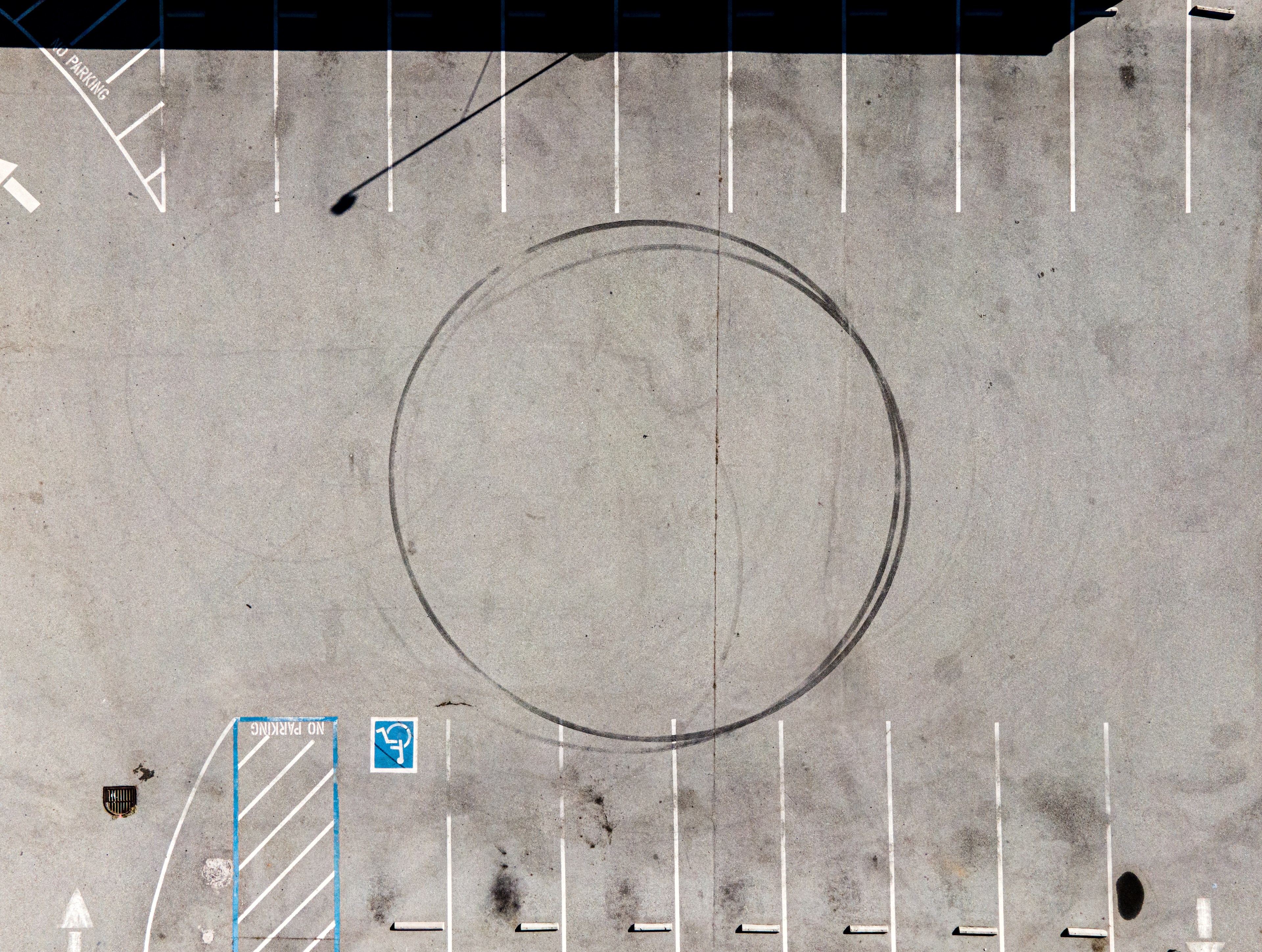 Aerial view of an empty parking lot with a large circular tire skid mark in the middle, numerous white parking lines, and a marked blue handicap spot.