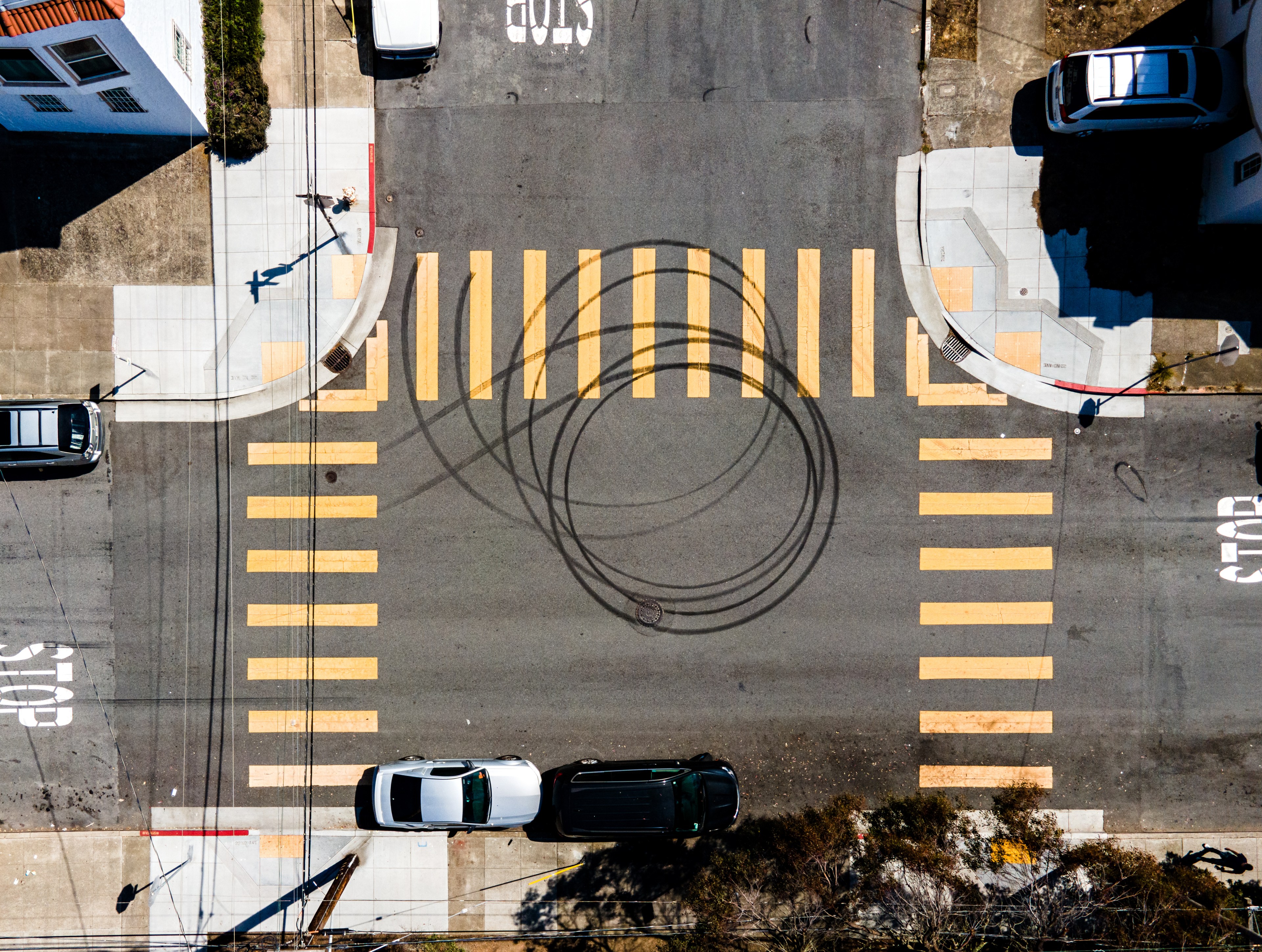 The image shows a road intersection with yellow crosswalks and "STOP" markings. Tire skid marks form circular patterns in the middle. Cars are parked nearby.